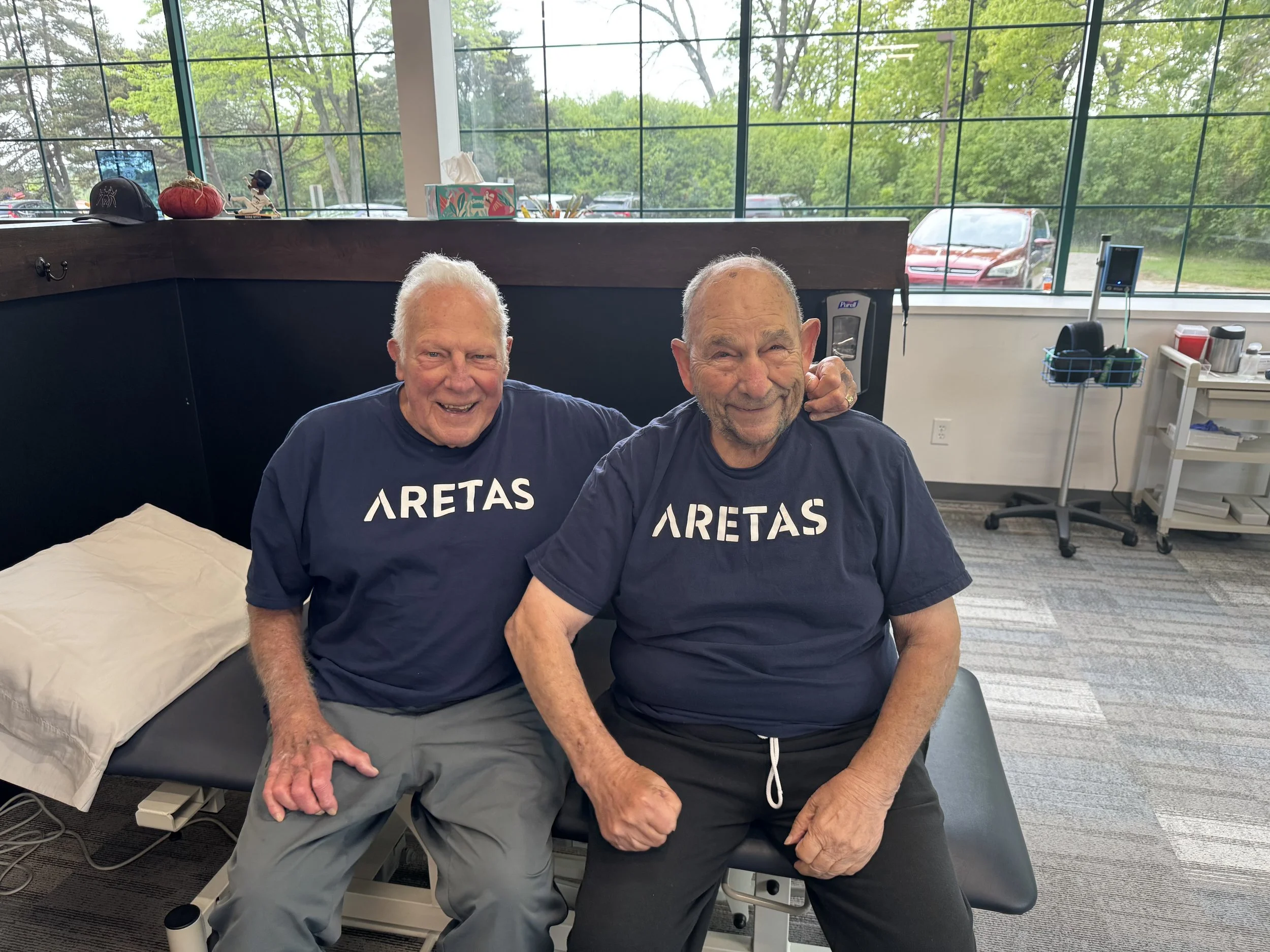 Two elderly men sitting on a medical examination table in a room with large windows. They are smiling and wearing matching navy blue shirts with 'ARETAS' printed on the front. One man has his arm around the other's shoulder.