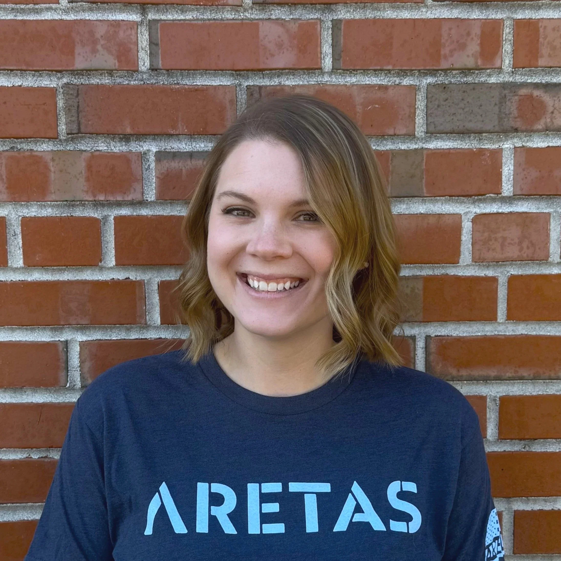 A young woman with collarbone length curly blond hair stands in front of a brick wall, wearing a navy Aretas shirt