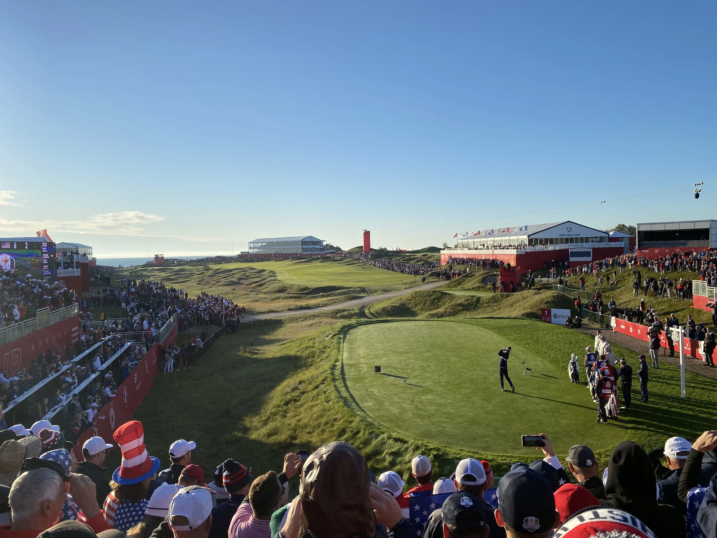 Crowd of spectators watching a golf match at a scenic course with a large television screen displaying scores and the landscape in the background.