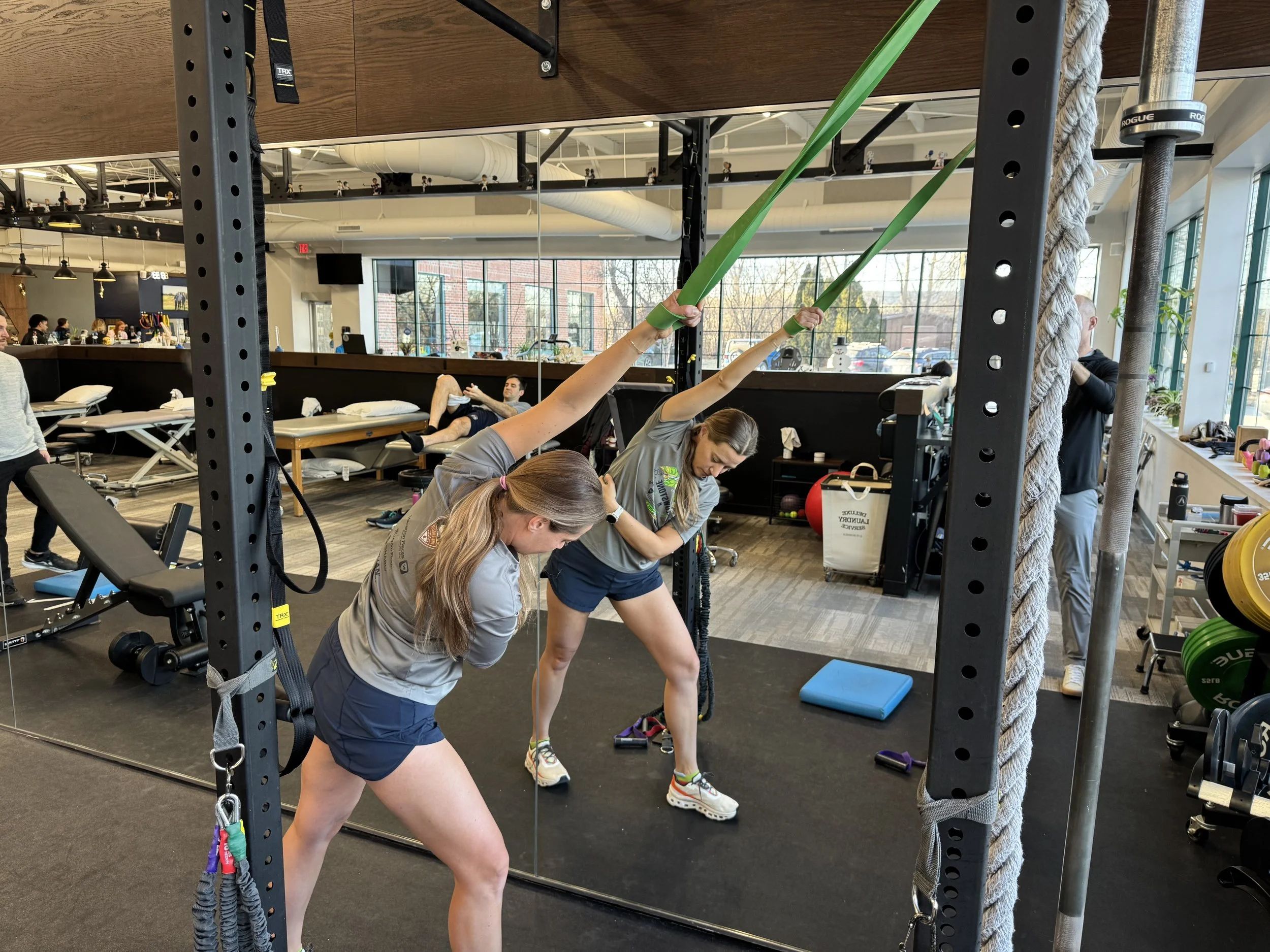 Two women exercising with resistance bands in a gym, mirroring exercises in front of a large mirror, with workout equipment and a lounge area visible in the background.