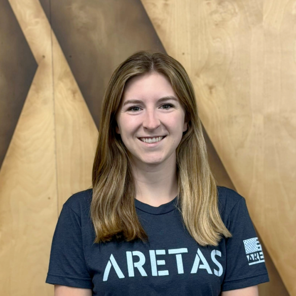 Young woman with long, wavy brown hair smiling, wearing a navy blue T-shirt with 'ARETAS' and a logo on the front, standing in front of a wooden wall background.