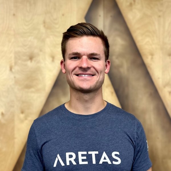 A smiling young man with short brown hair wearing a navy blue t-shirt with 'ARETAS' printed on it, standing in front of a wooden wall with geometric patterns.