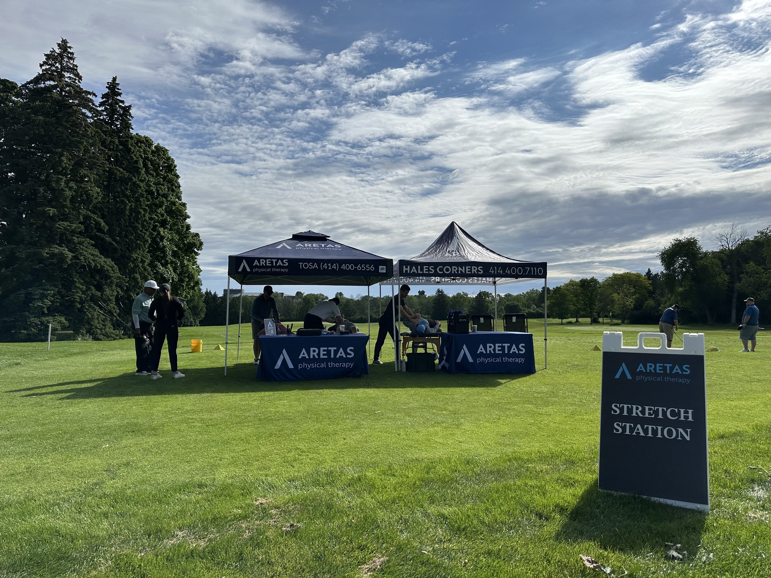Outdoor stretch station set up with two tents and people receiving physical therapy treatments on a golf course under a partly cloudy sky.
