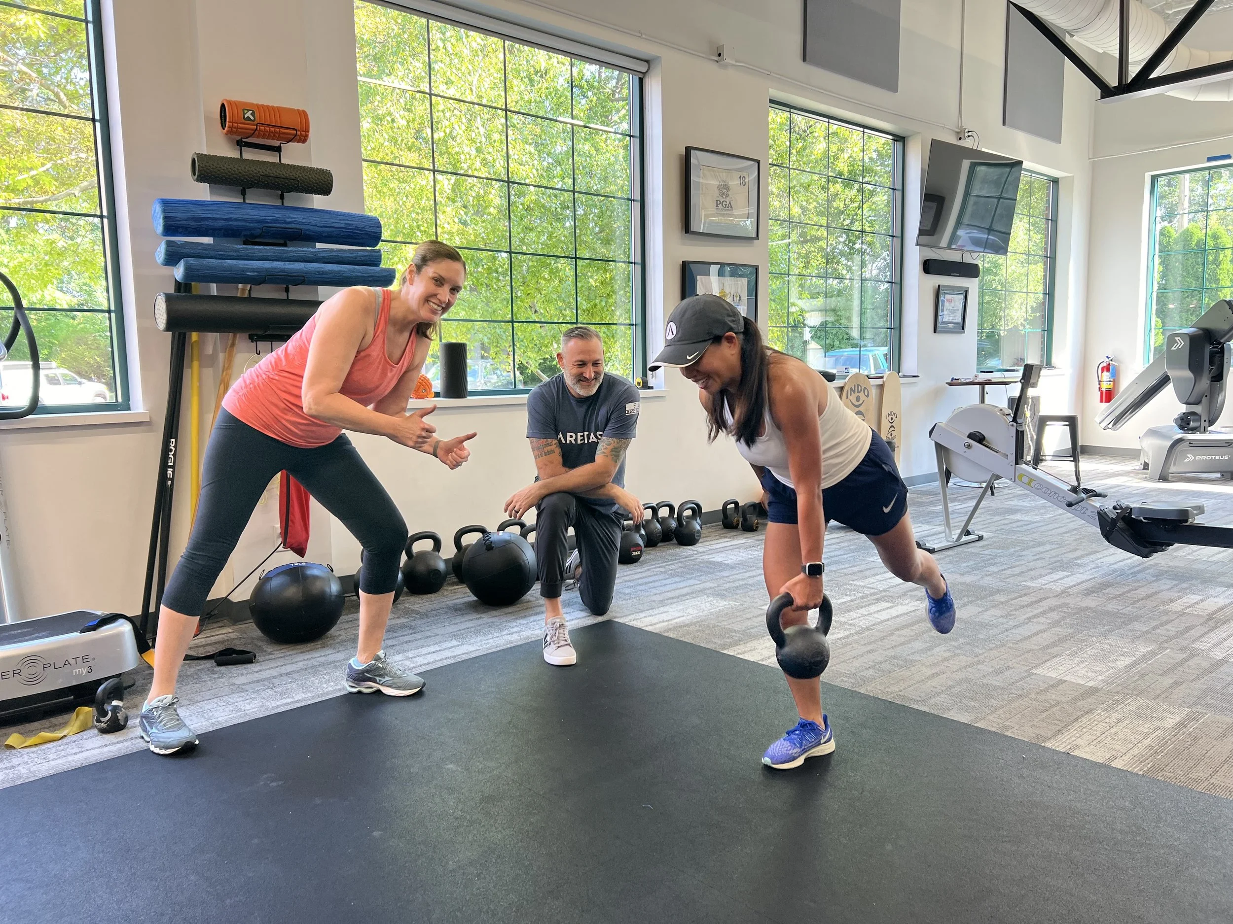 Two women and one man grouped together in a gym. One woman is looking at the camera smiling and giving a thumbs up. The man is kneeling on one knee overseeing an exercise. One woman is doing a kettle bell exercise.
