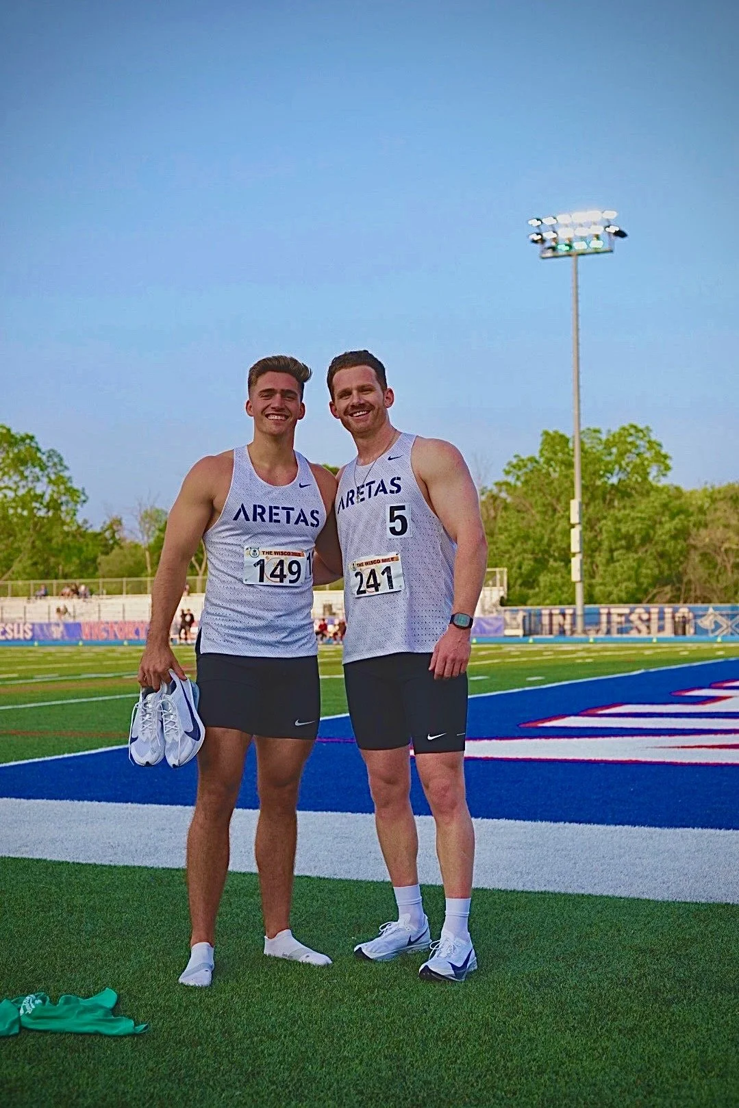 Two male athletes in white and black running uniforms standing on a track, smiling for a photo after a race. One holds a pair of running shoes, and both wear race bibs. The background shows a track, some trees, and a bright blue sky.