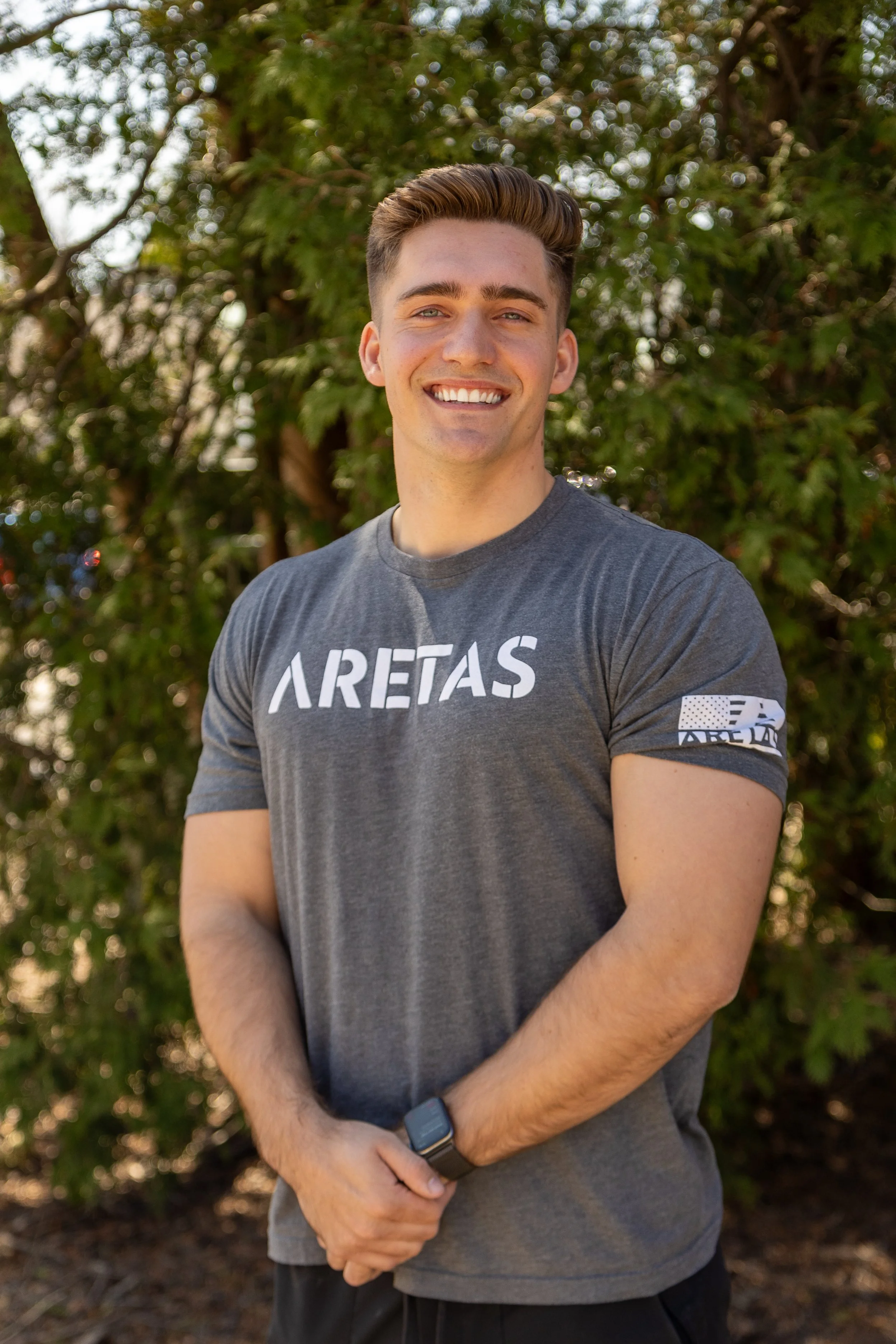 A young man with short brown hair smiling outdoors in front of green trees, wearing a gray T-shirt with 'ARETAS' written on it and an American flag patch on the sleeve.