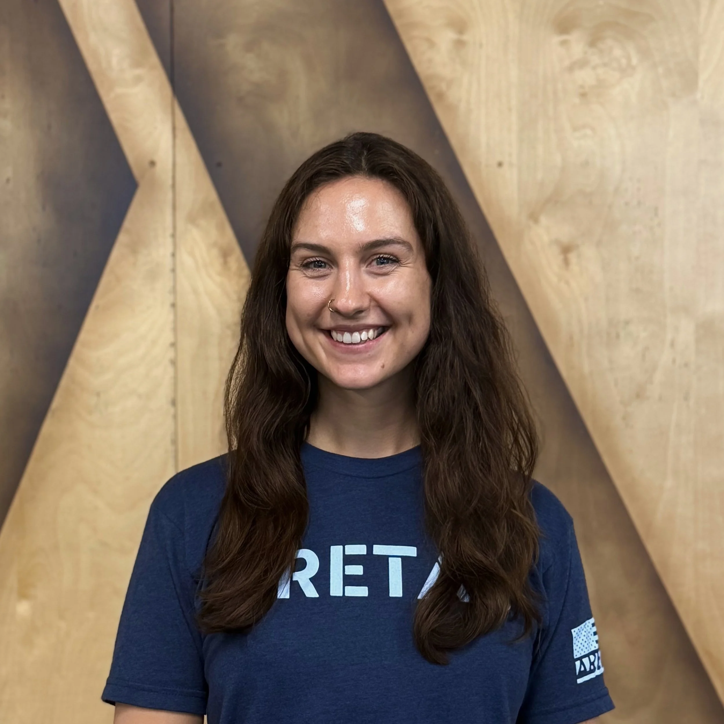 Young woman with long wavy brown hair smiling, wearing a blue t-shirt with partial text, standing in front of a background with large wooden geometric shapes.