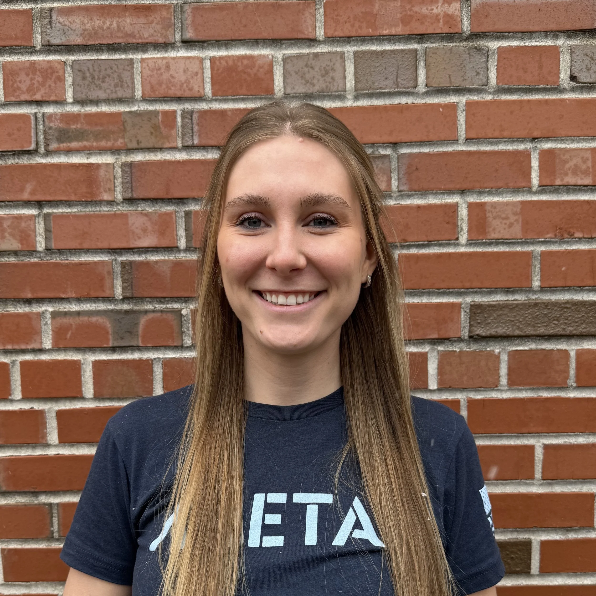 A young woman with long straight blond hair stands in front of a brick wall, wearing a navy Aretas shirt
