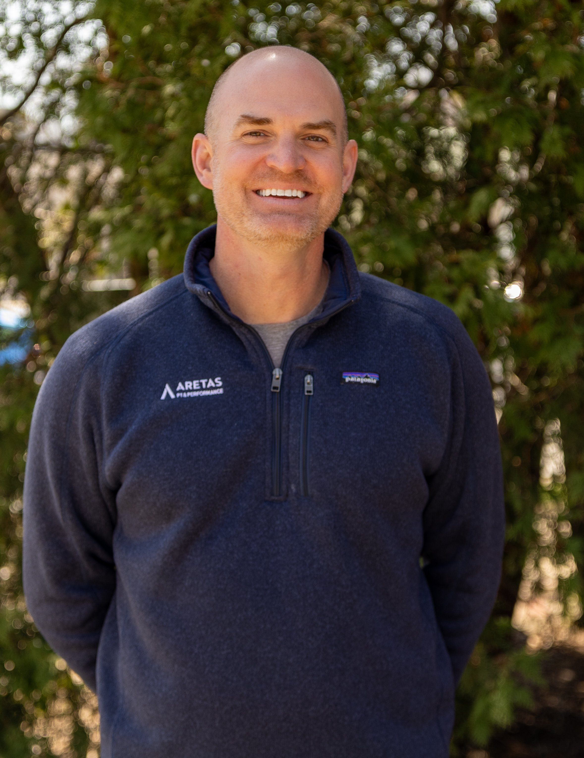 A man with a shaved head and light beard smiling outdoors, wearing a dark blue Patagonia fleece jacket with 'ARETAS PERFORMANCE' logo on the left chest, standing in front of green foliage.