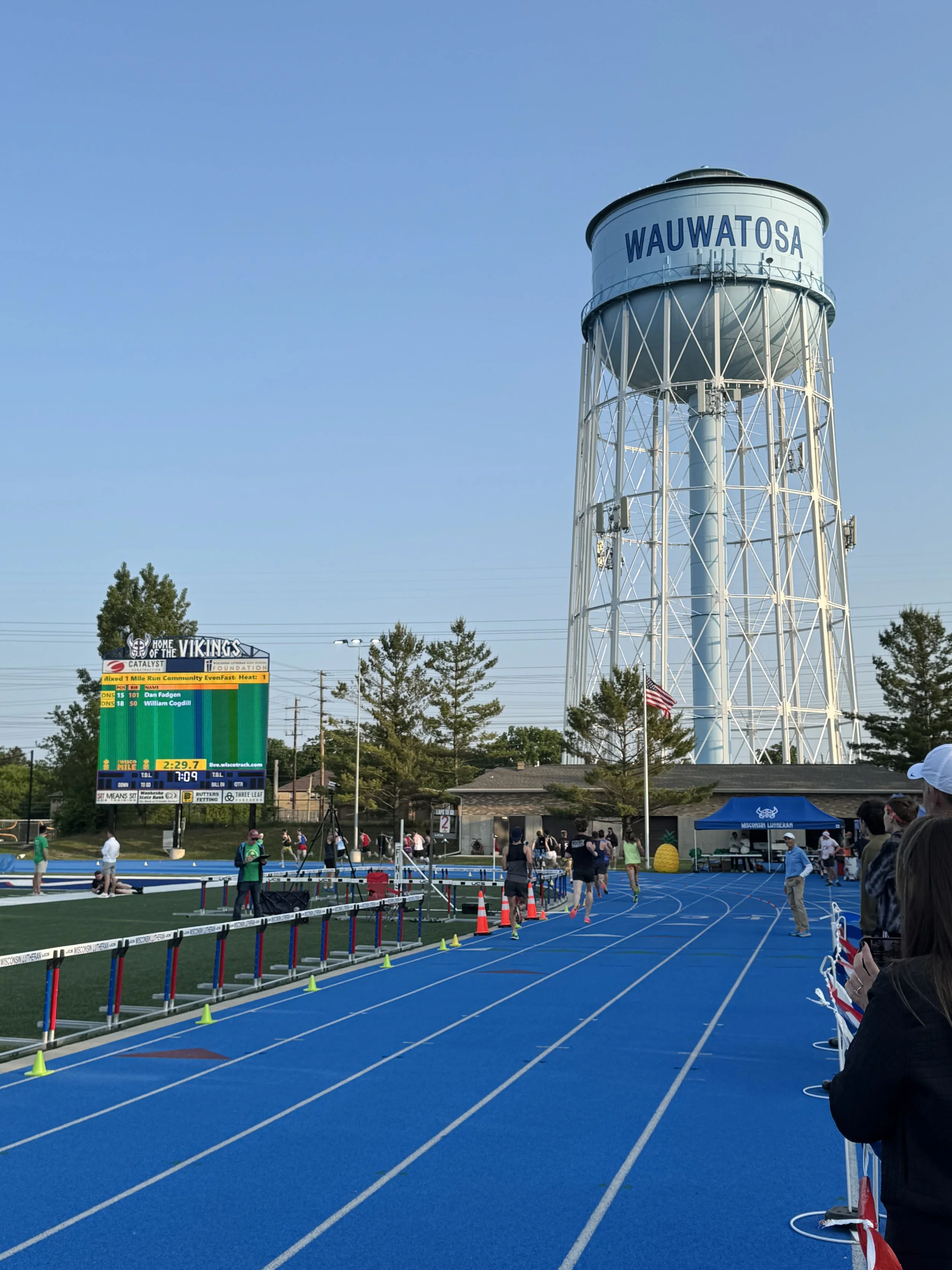 Track and field stadium with runners, bleachers, a digital scoreboard, a water tower labeled 'Wauwatosa', and a blue track surface, during a sporting event.