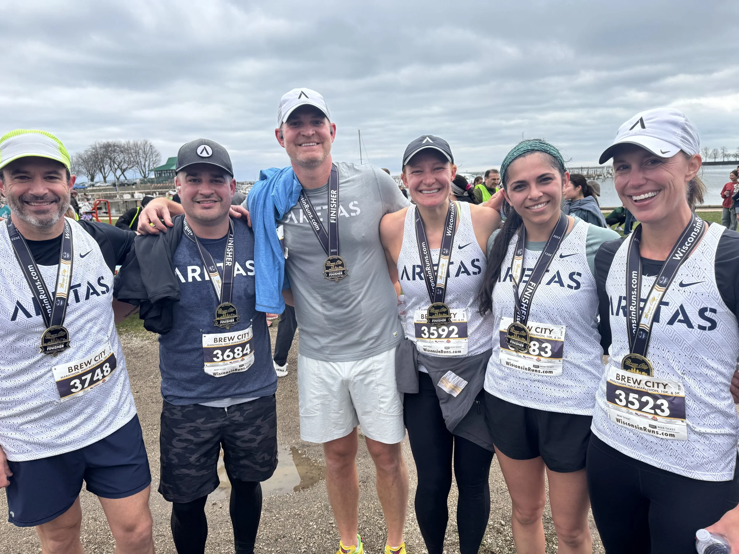 Group of six runners after a race, wearing race bibs and medals, standing outdoors near a body of water with a cloudy sky.