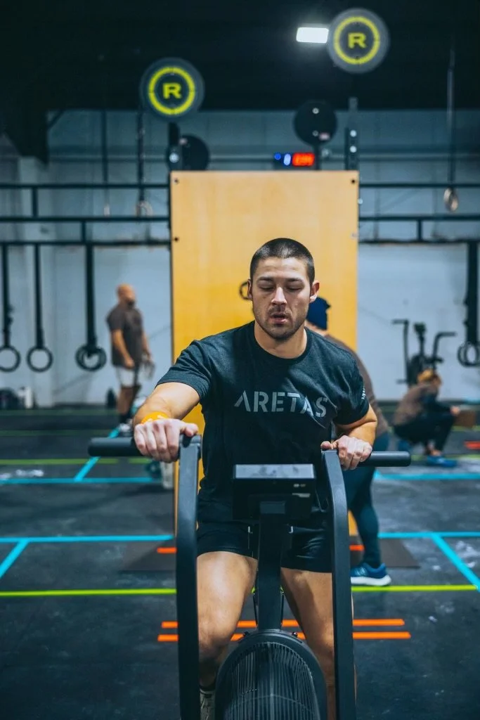 Man working out in a gym on a stationary out in a t-shirt that says "Aretas"