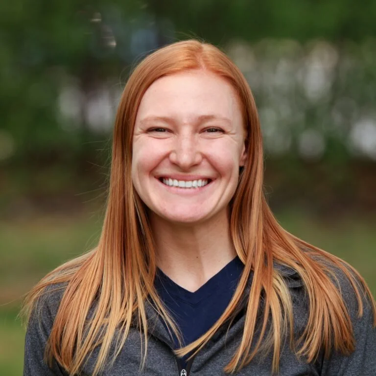 A smiling woman with long red hair outdoors in a blurred natural background.