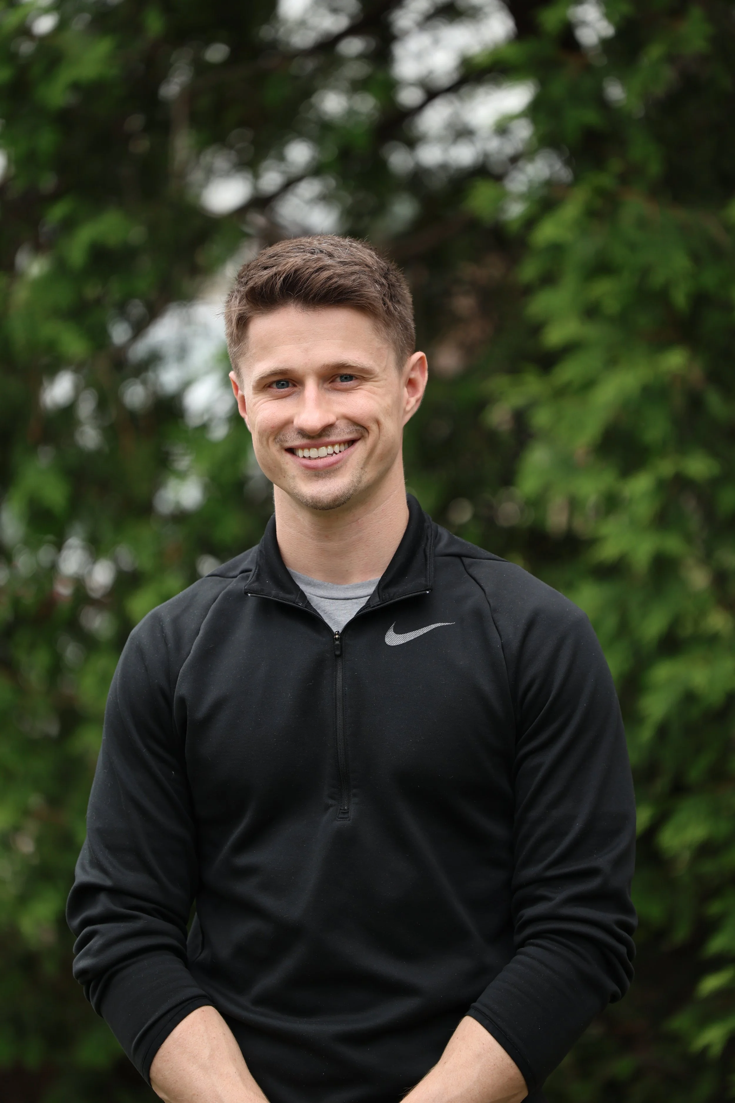 A smiling man with short brown hair wearing a black Nike zip-up jacket stands outdoors against a background of green trees.