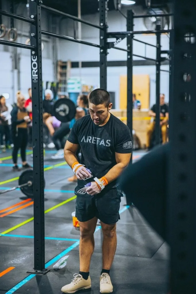 A man in a gym preparing for a workout, wearing a black t-shirt and shorts, with gym equipment and other people in the background.