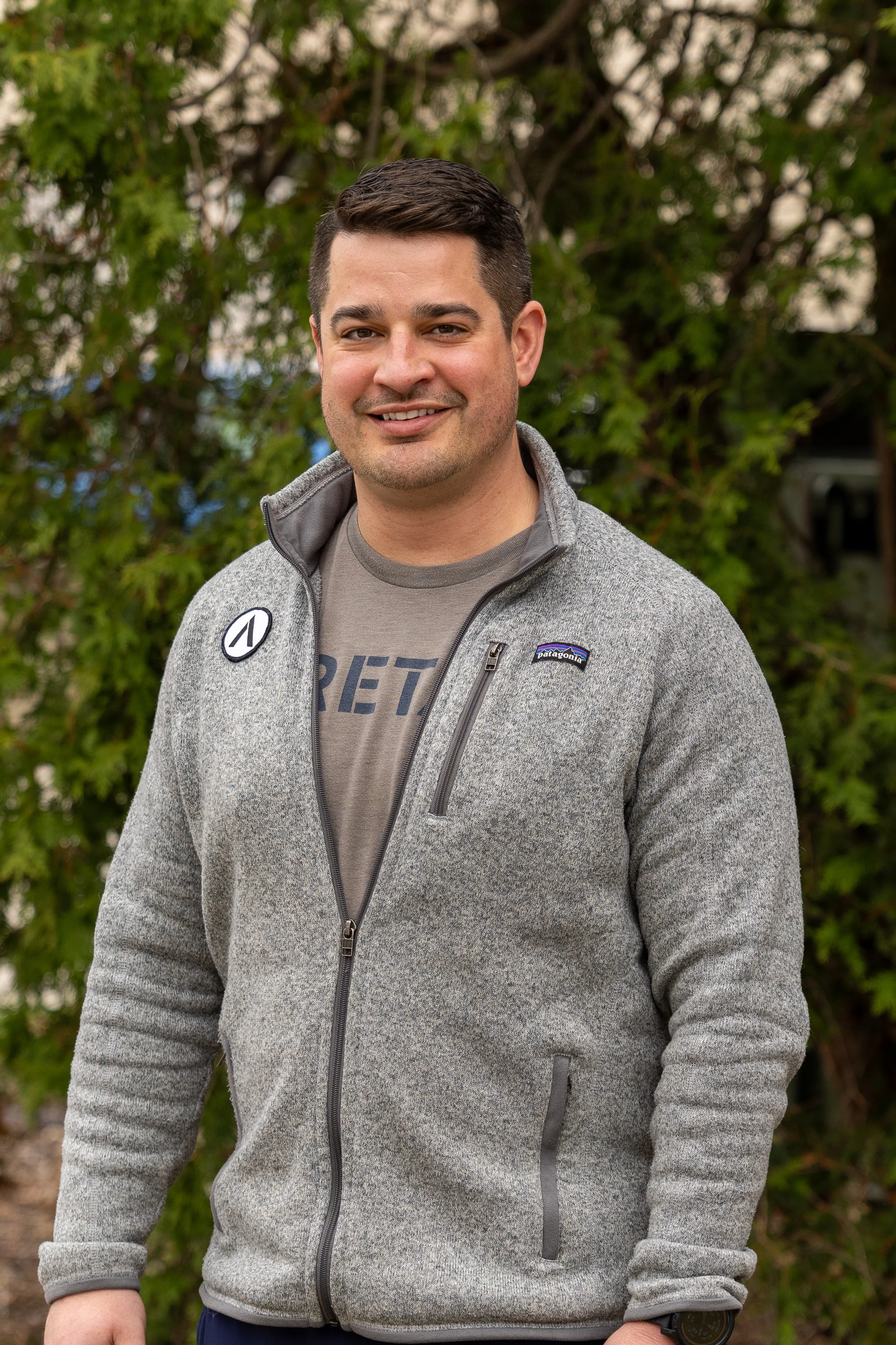 A smiling man with short dark hair standing outdoors in front of green foliage, wearing a gray zip-up jacket with patches and a gray T-shirt.