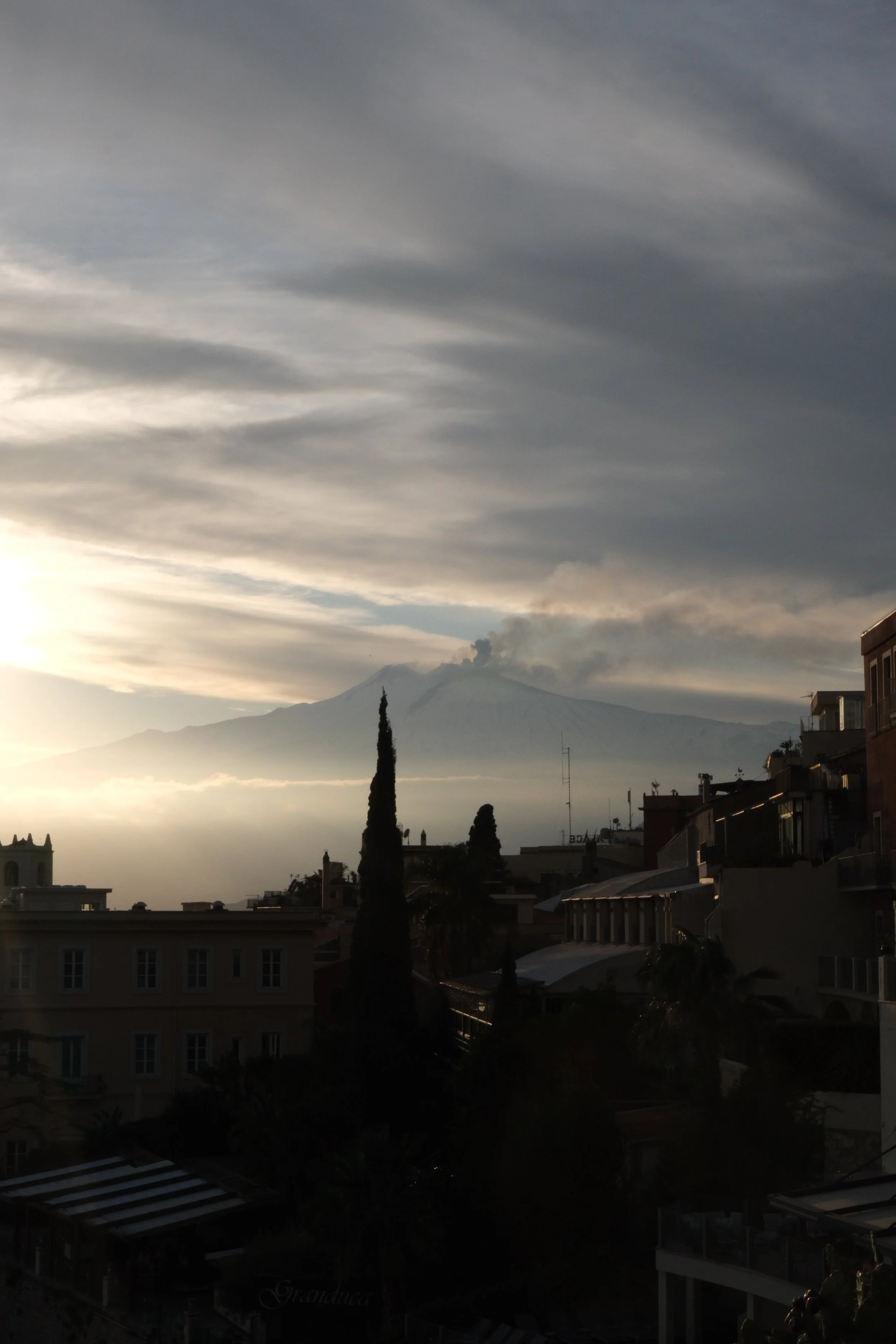 Sunset over a city with a view of a volcano erupting in the distance, smoke billowing from the volcano's peak, and clouds in the sky.