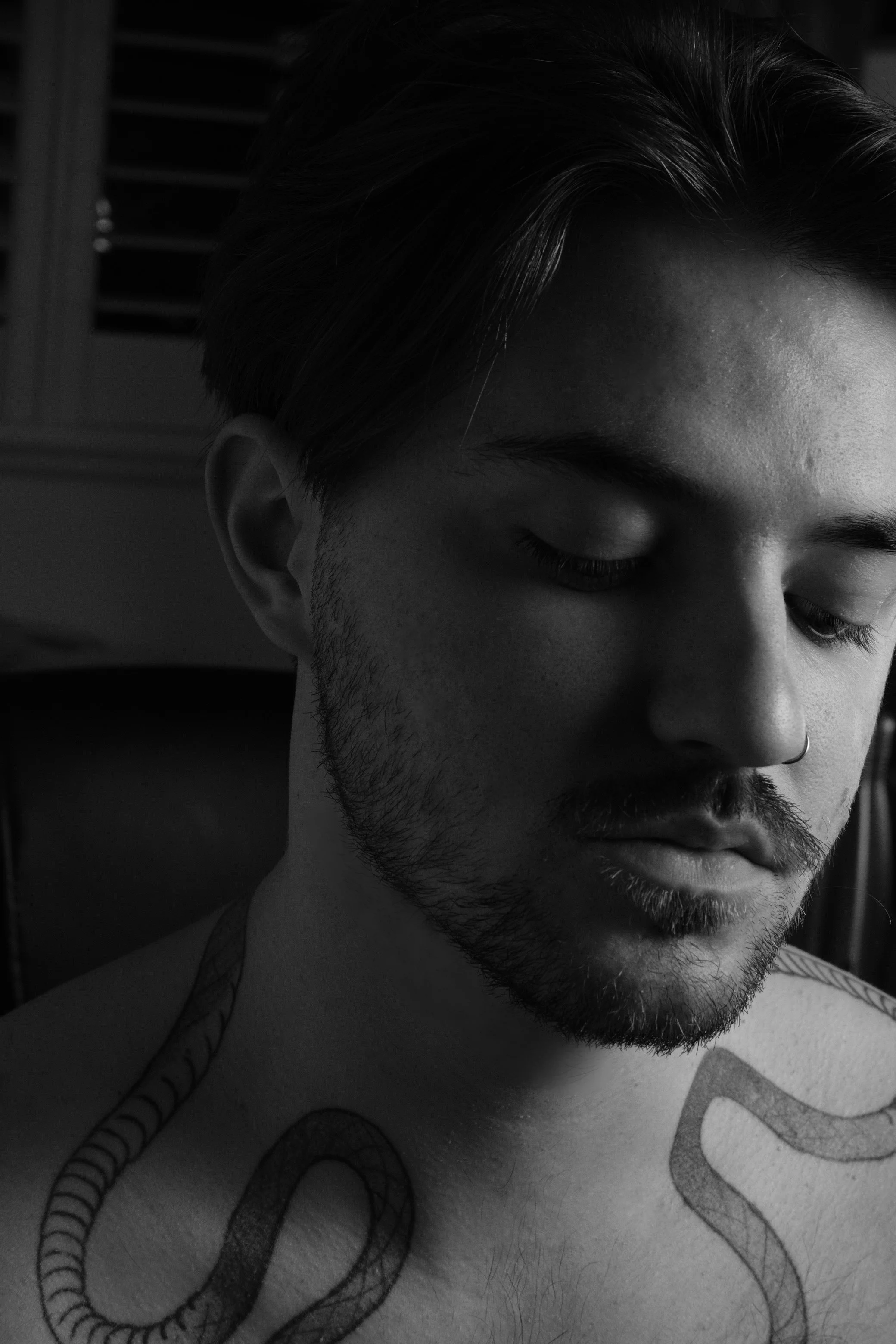 A black and white close-up photo of a young man with closed eyes, short hair, a mustache, and a beard. He has a tattoo of a snake on his shoulder.