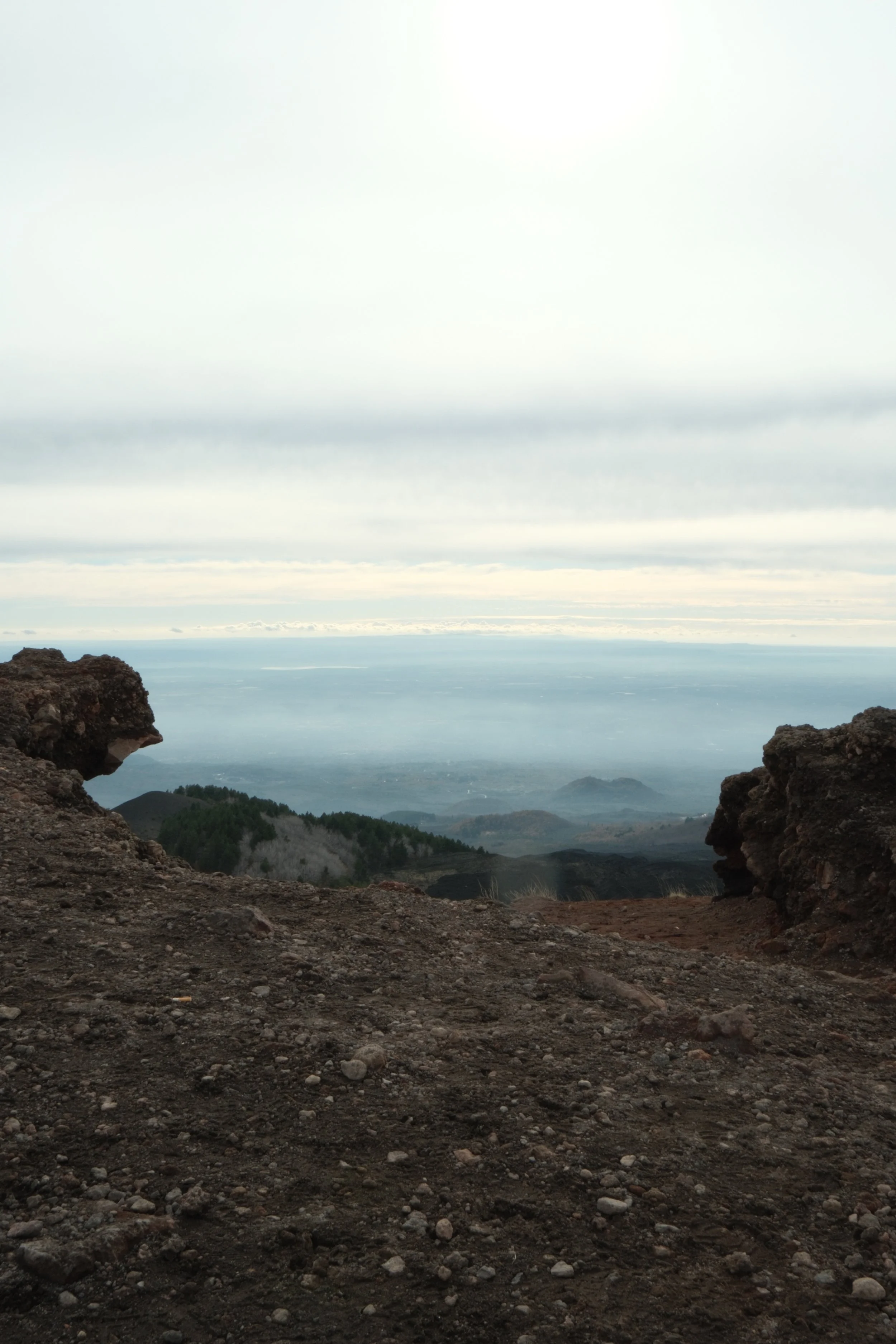 A volcanic crater with rocky edges, overlooking a landscape with green forests, hills, and a cloudy sky.
