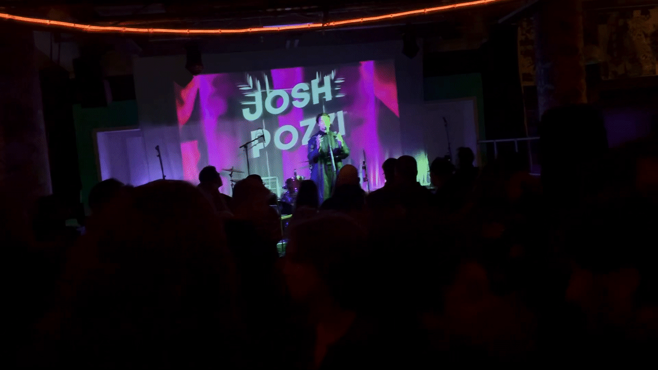 A performer stands on stage with a microphone, illuminated by colorful stage lights, with an audience watching.