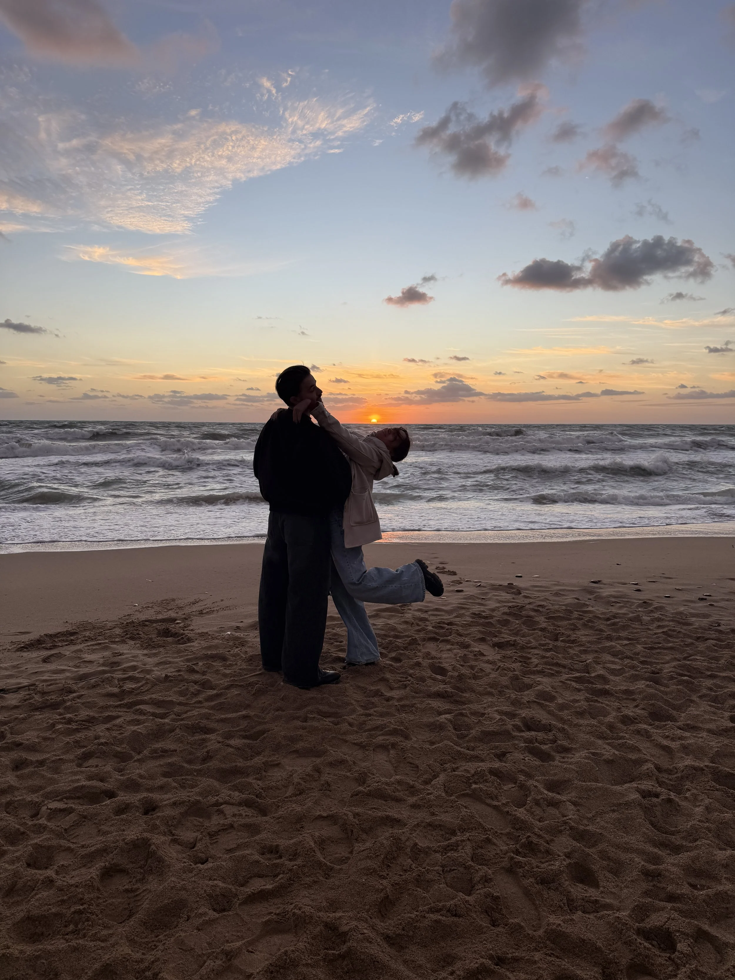 Two people dancing on a beach at sunset with clouds in the sky.