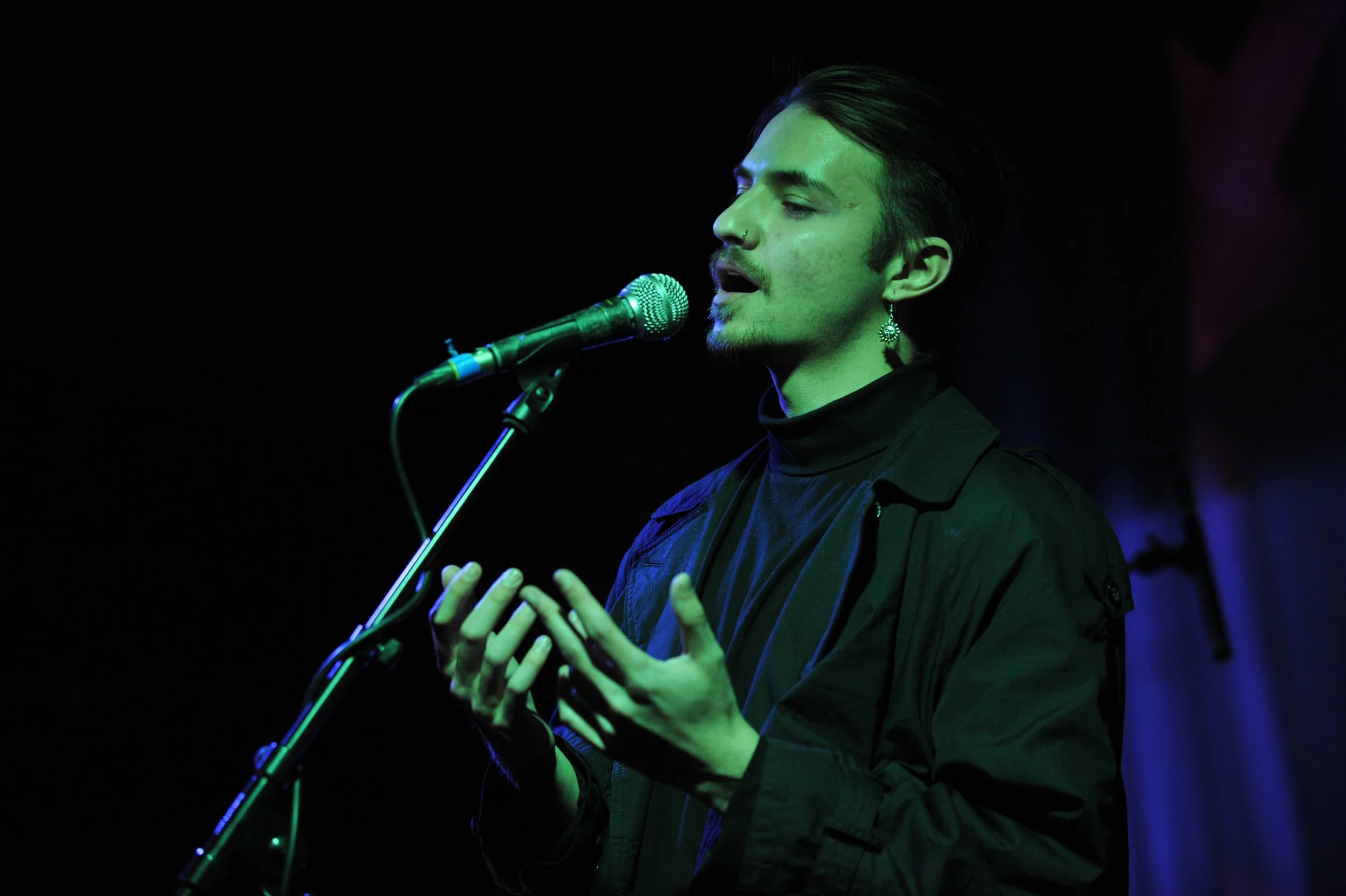 A person singing into a microphone on stage, wearing a black jacket and earrings, with a dark background and stage lighting.
