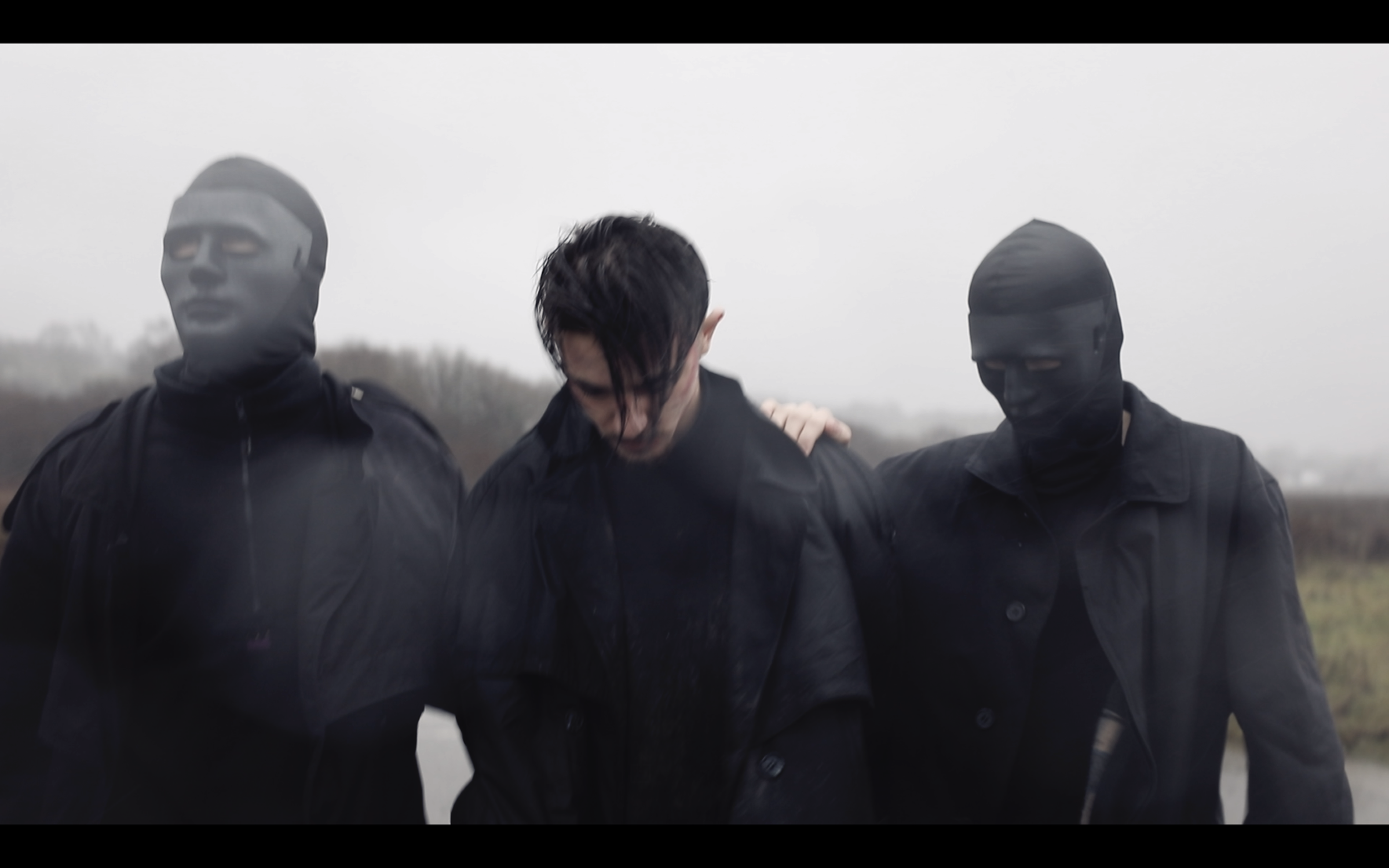 A person supported by two masked individuals in a bleak outdoor setting, possibly during rain or mist.