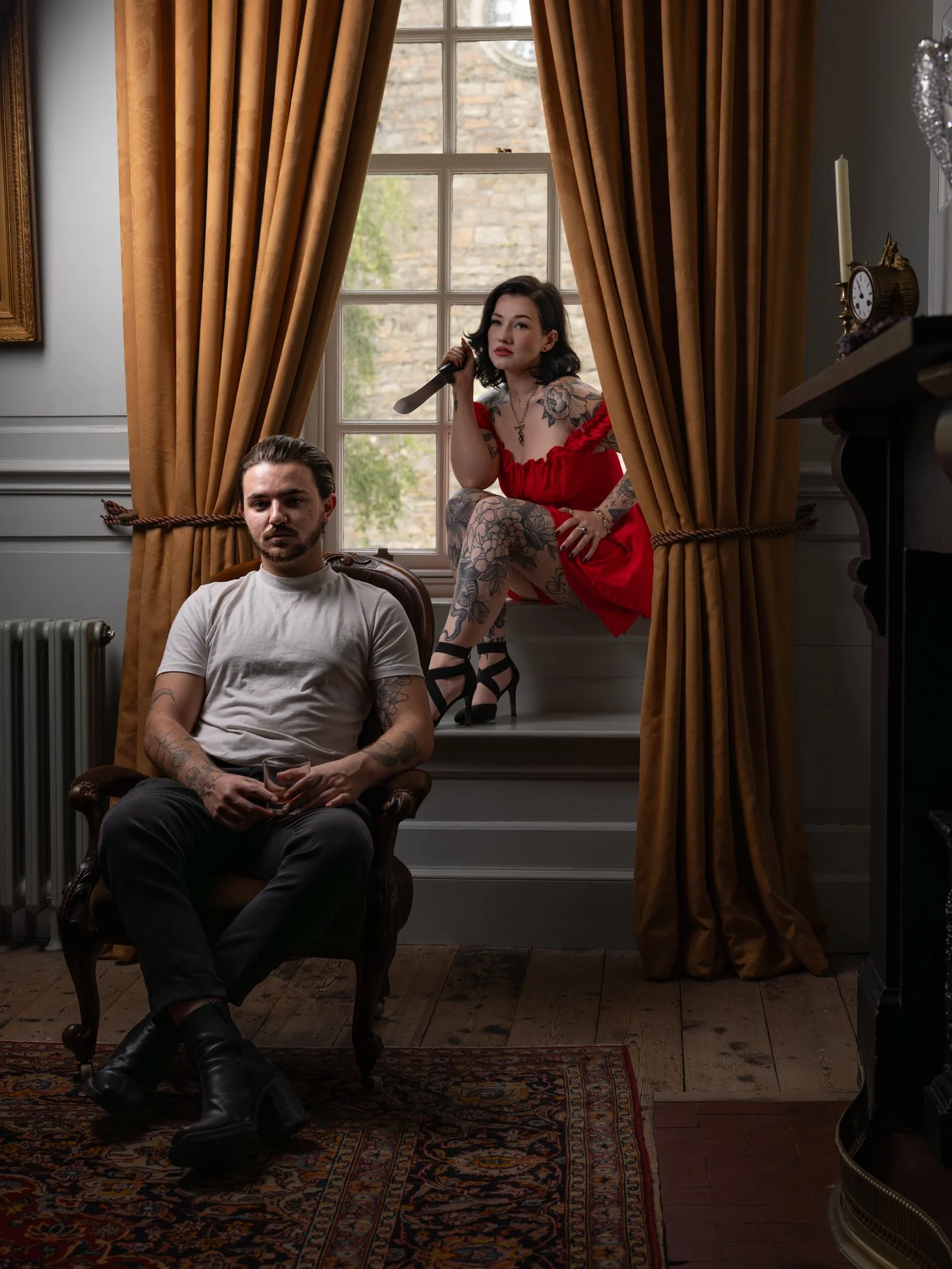 A man sitting in a vintage armchair holding a drink, and a woman sitting on a windowsill wearing a red dress and high heels, holding a knife, in a room with golden curtains and a wooden floor.