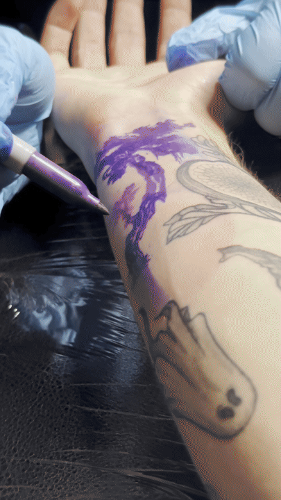 Person getting a tattoo of a purple tree on their forearm, with a tattoo artist using a tattoo machine and wearing blue gloves.