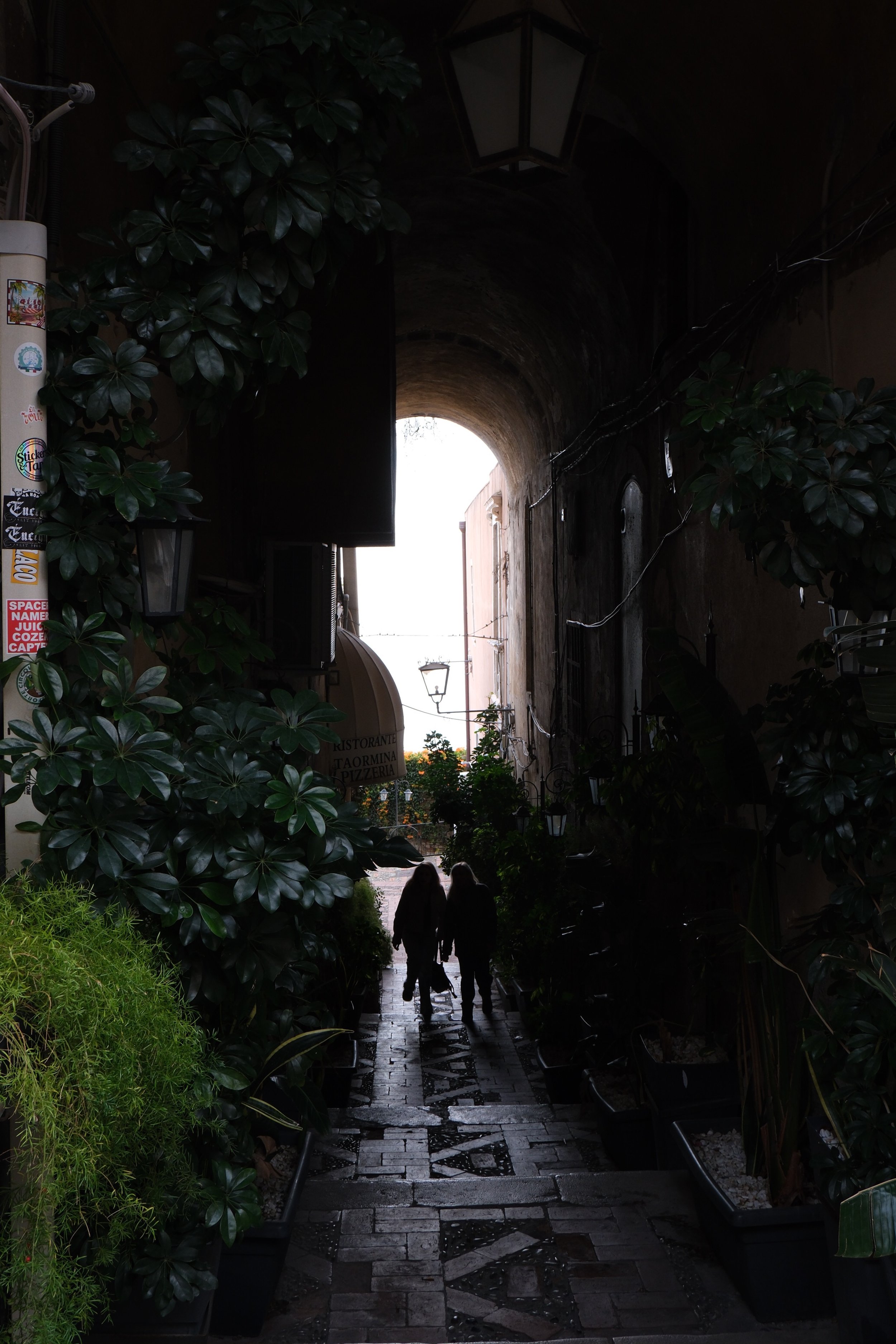 Silhouettes of two people walk hand-in-hand through a narrow, cobblestone alleyway with lush greenery and potted plants on either side, leading towards a bright, sunlit opening with an arch.