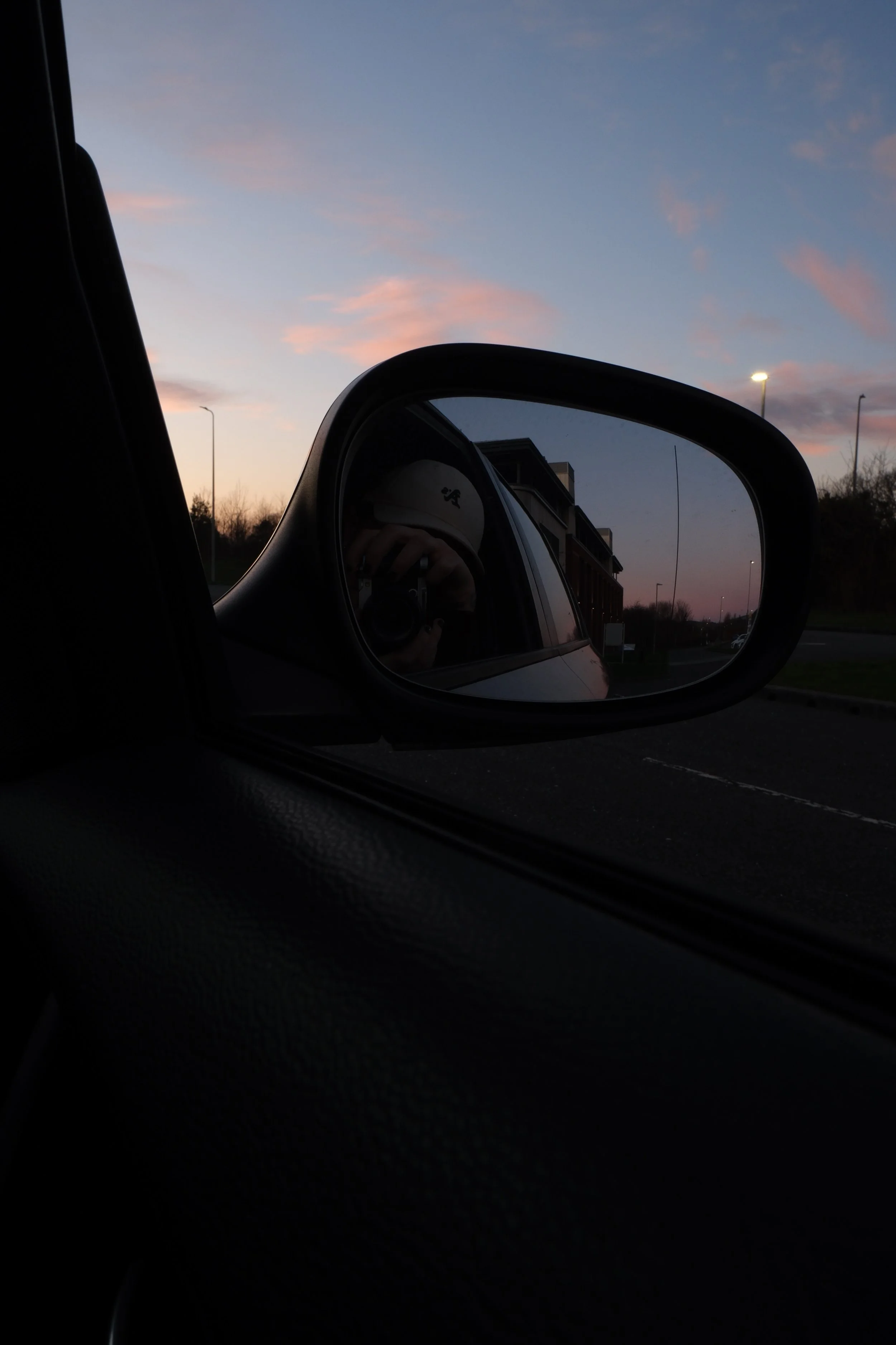 view of a sunset sky with pink and blue clouds, seen through a car's side mirror, with a partial reflection of the photographer holding a camera.