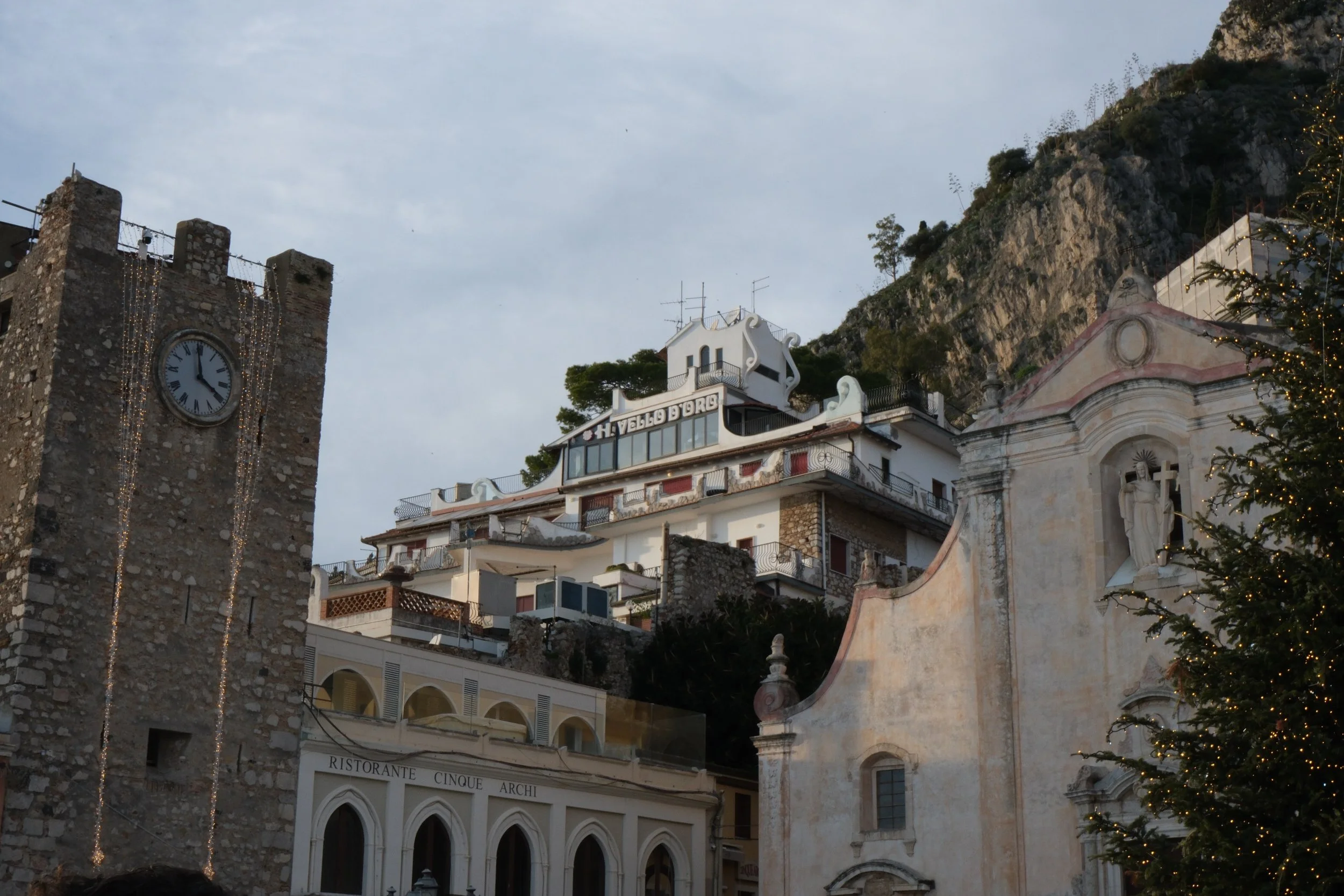 Coastal village with a stone clock tower, a hillside building with the sign 'Vello D'oro,' a church with a statue, and a Christmas tree with lights.