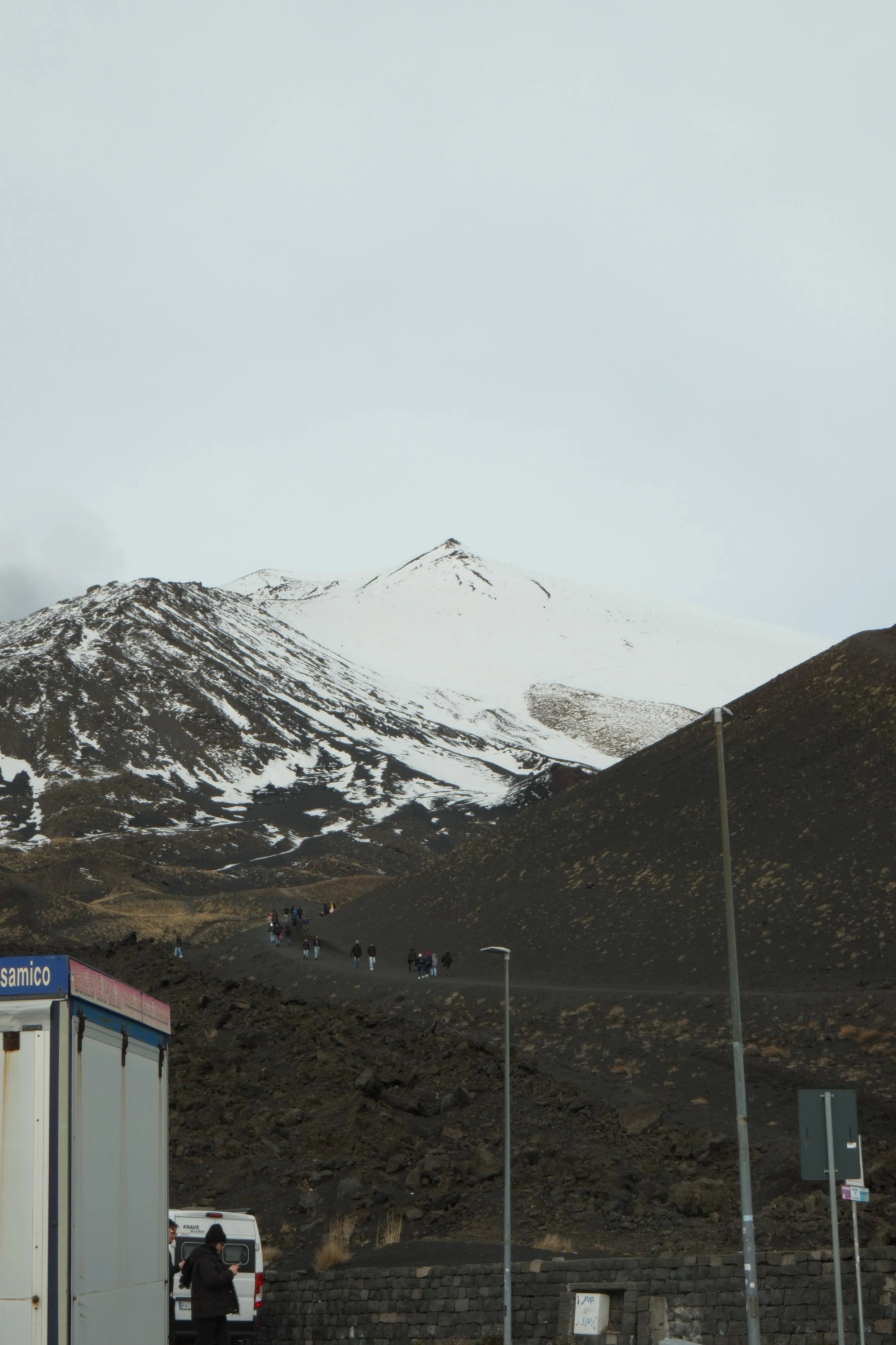 Snow-capped mountain with dark volcanic terrain and a group of people walking up a trail, with a partly cloudy sky in the background.