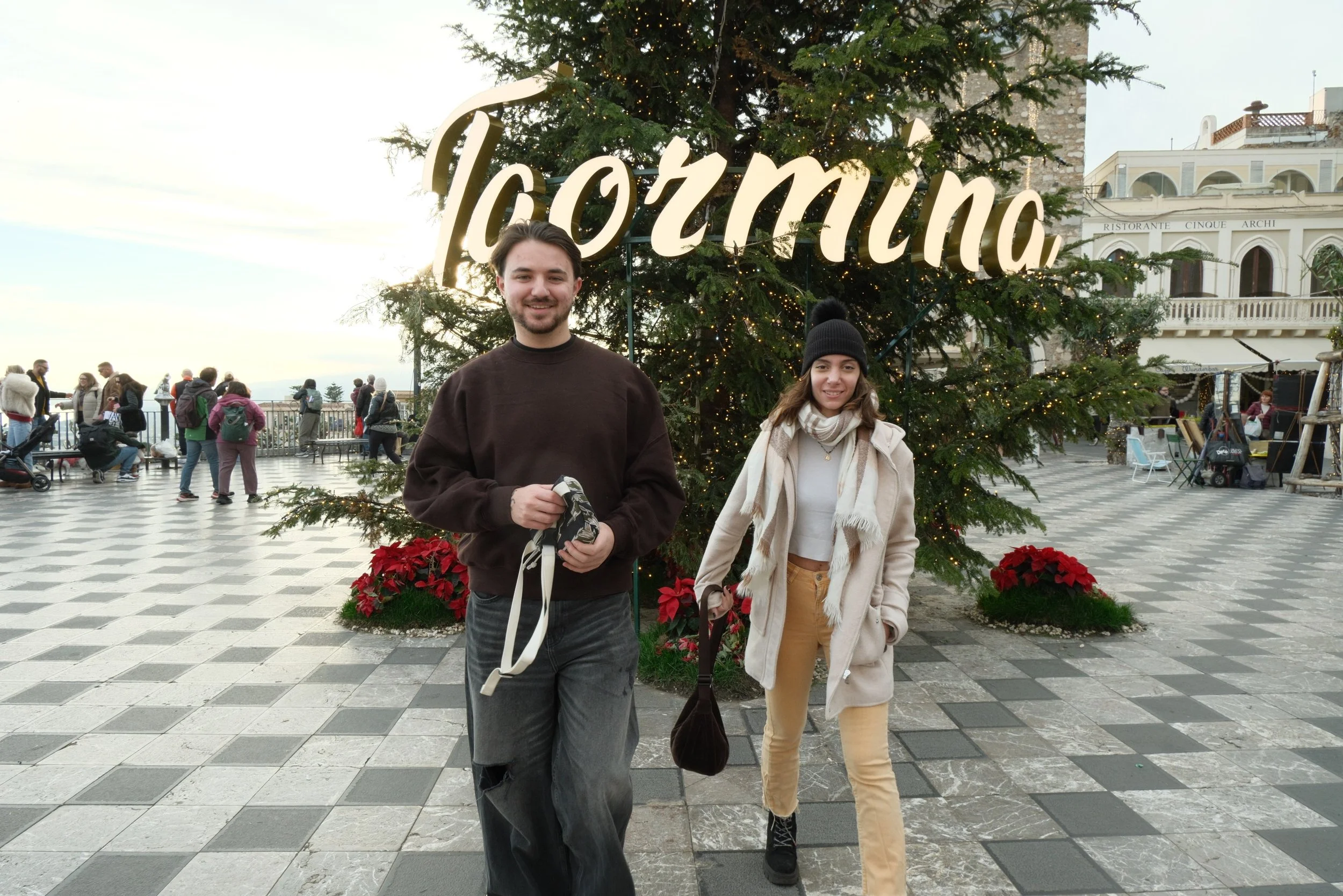 Two young adults, a man and a woman, standing in front of a decorated Christmas tree with the word "TORMINA" illuminated at the top, on a checkered public square with other people in the background, during daytime.