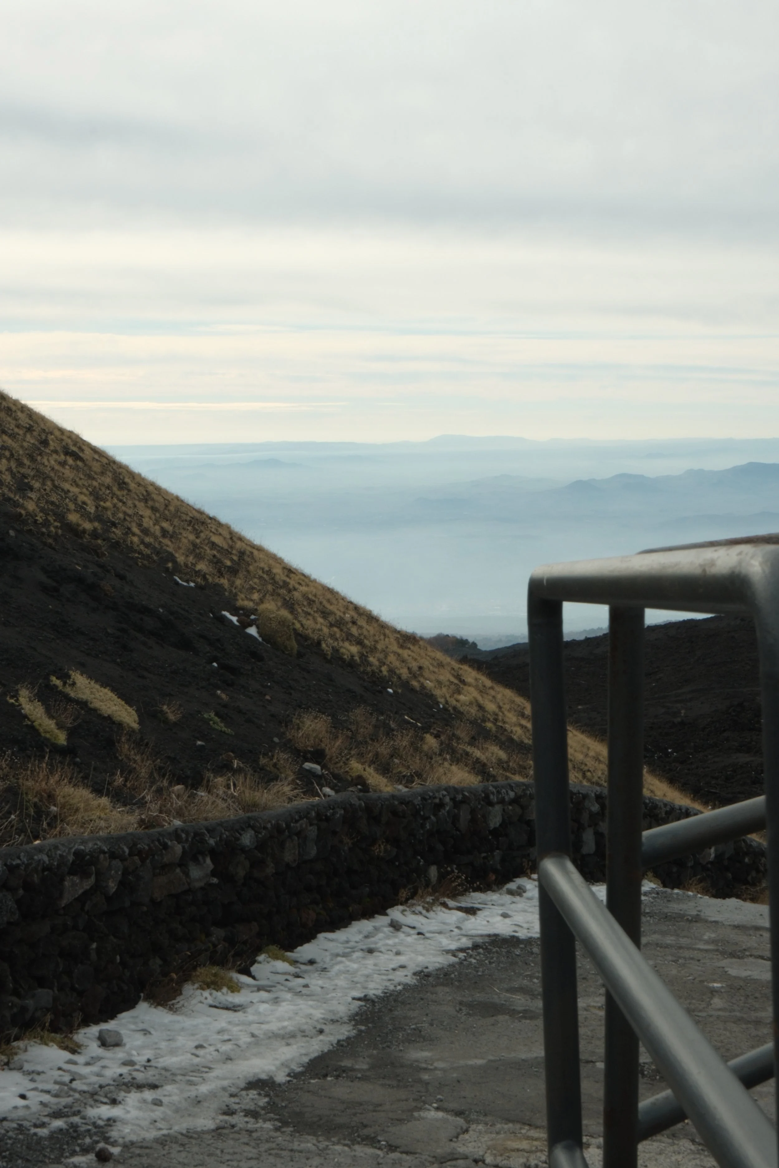 View from a mountain trail with a metal railing, rocky path, and a landscape of mountains and cloudy sky in the distance.