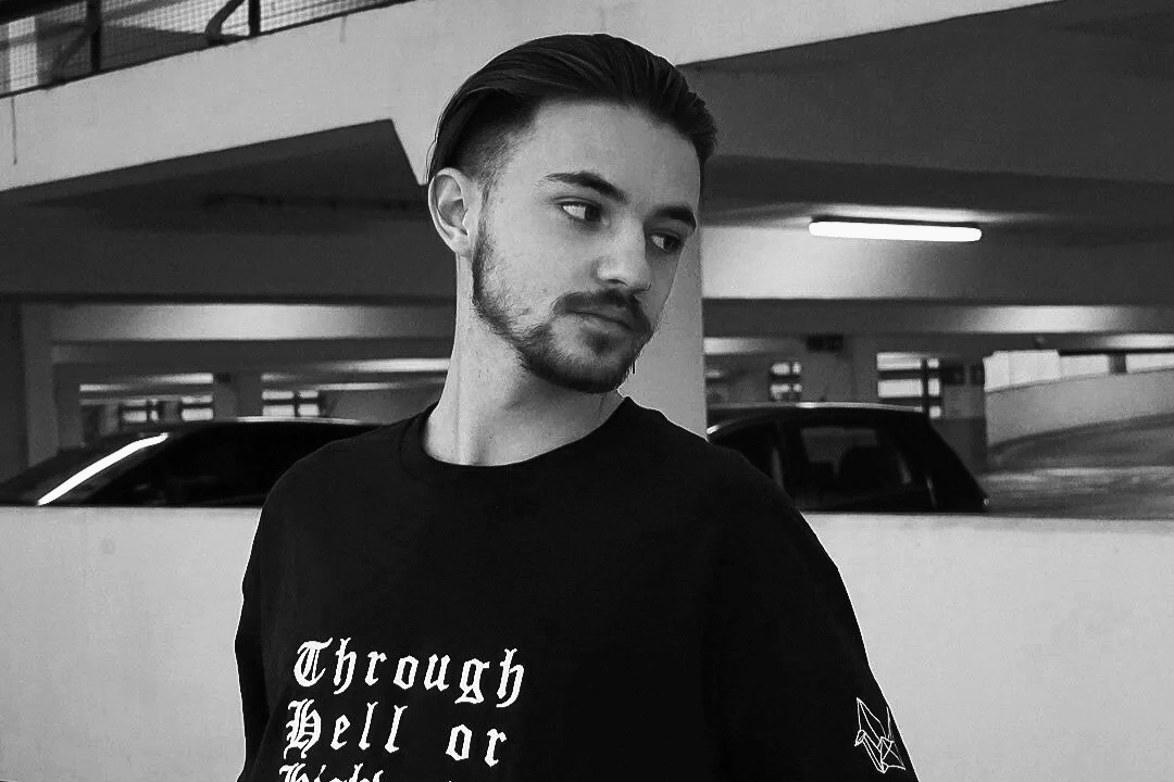 A young man with styled hair and a beard standing in a parking garage, wearing a black t-shirt with white text that reads "Through Hell or" and a small graphic on the sleeve.