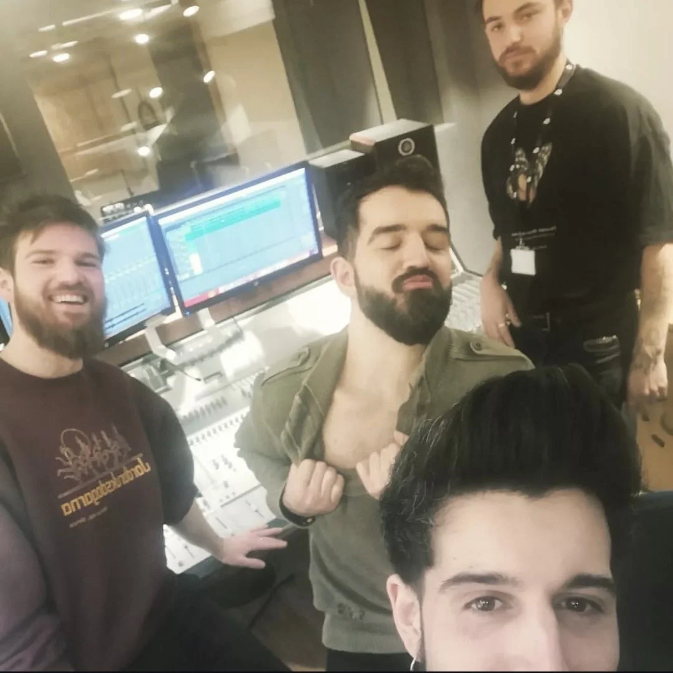 Four men in a recording studio near computer monitors. One man is taking a selfie while three others smile or make playful gestures. The studio environment includes audio equipment and soundproof windows.