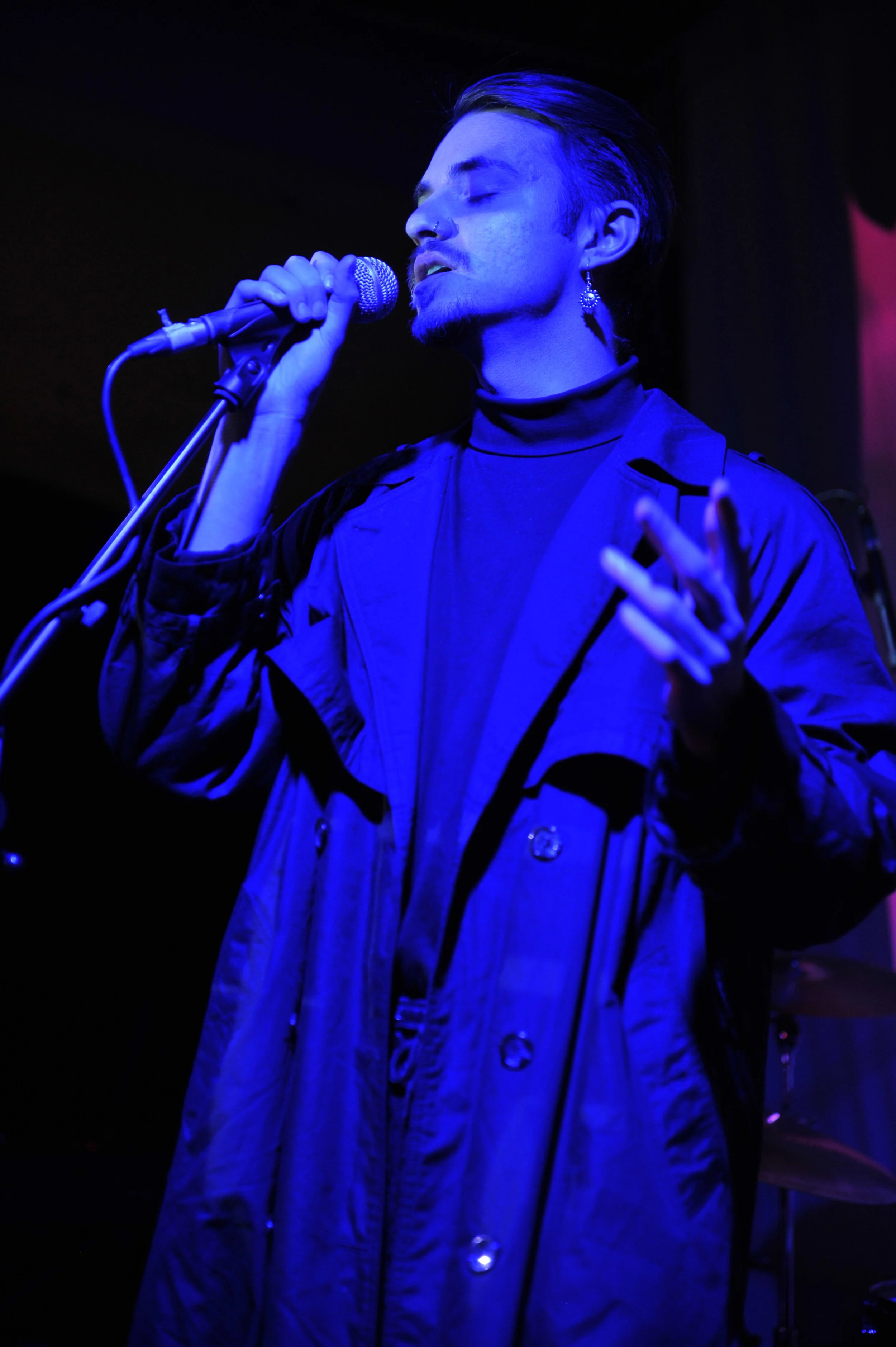 A person singing into a microphone with blue stage lighting, wearing a black jacket and earrings.