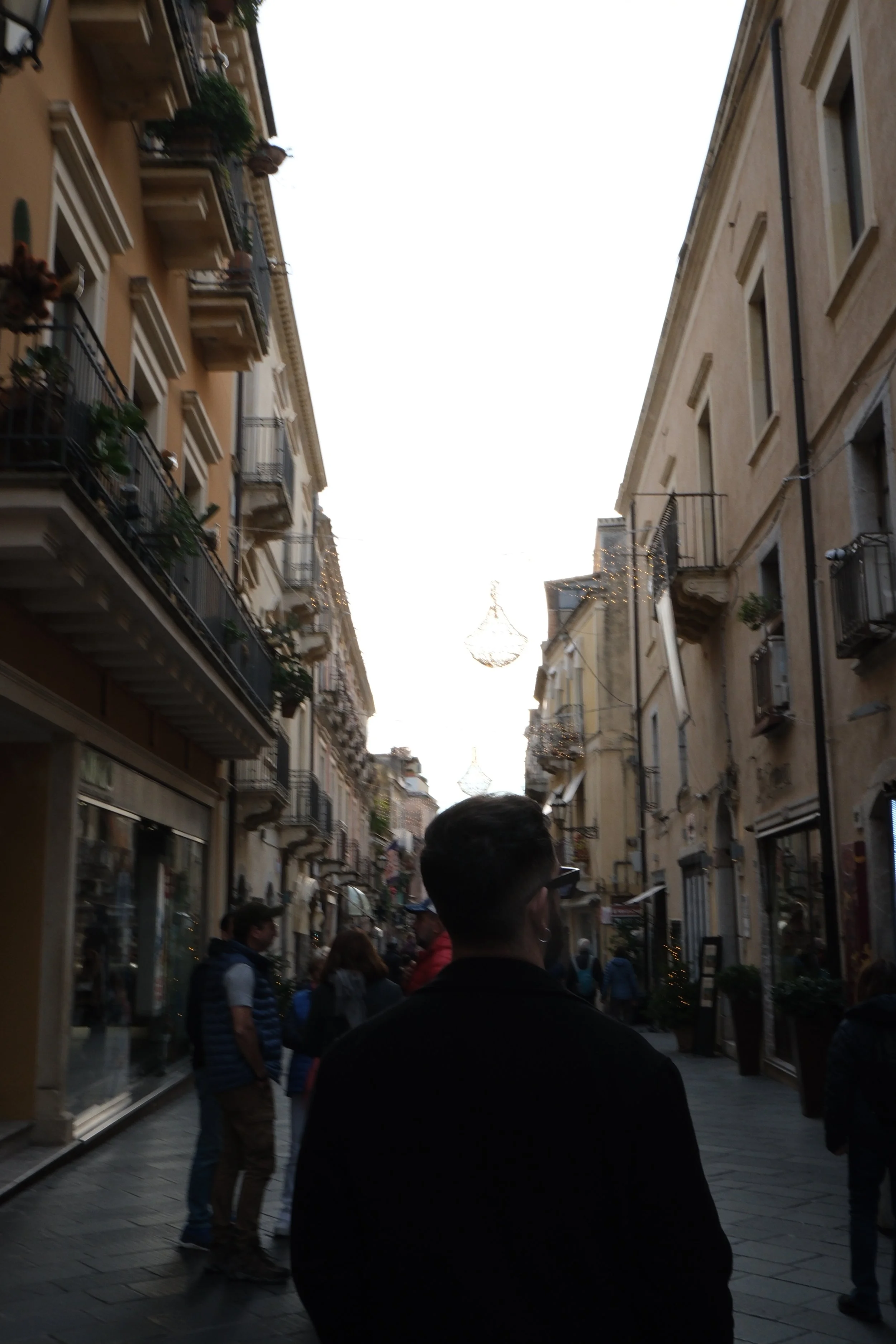 A street view in a European city with people walking, narrow buildings with balconies, and festive lights hanging across the street.