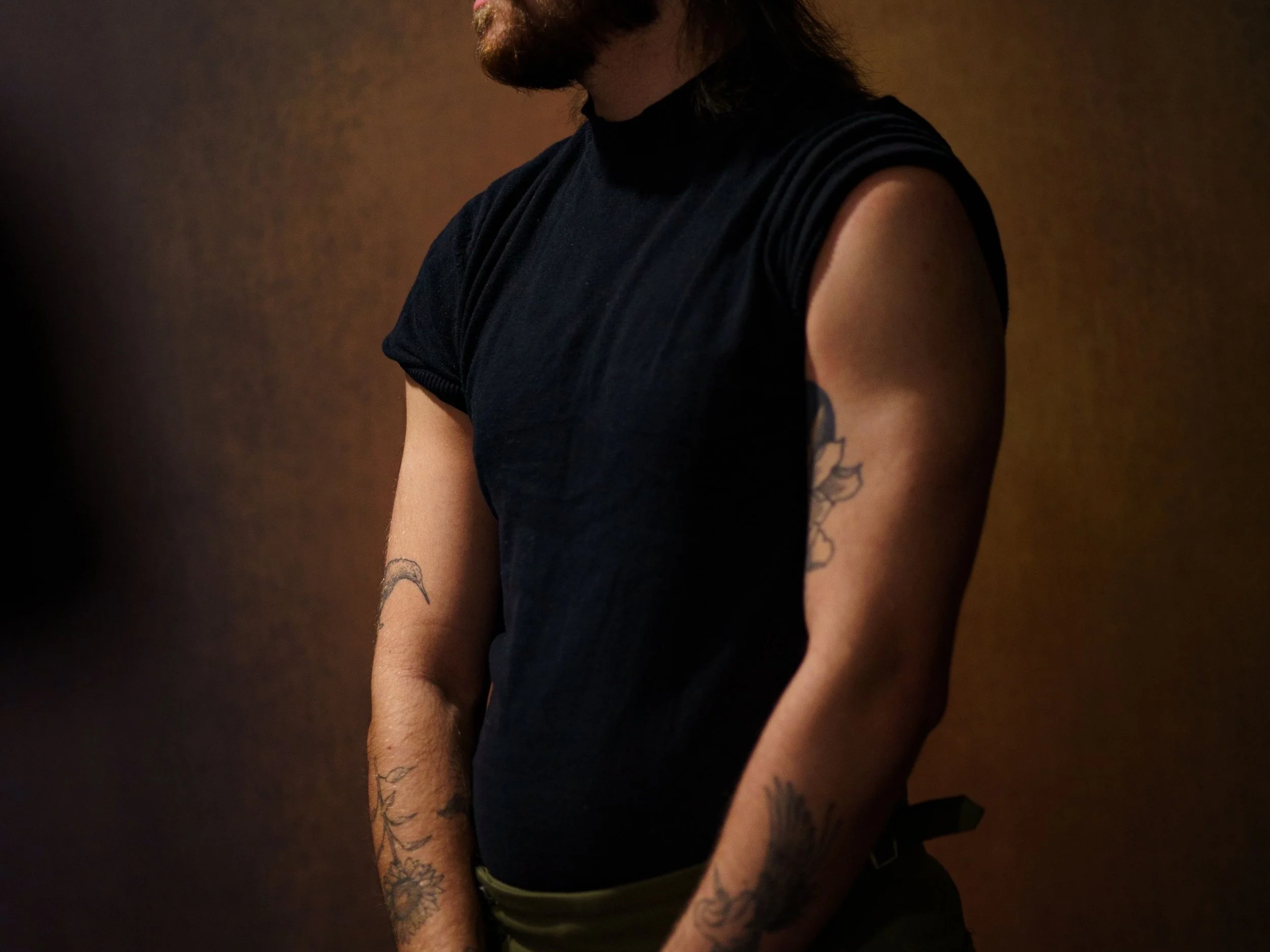 Partial view of a person with tattoos on arms, wearing a black sleeveless shirt, against a brown background.