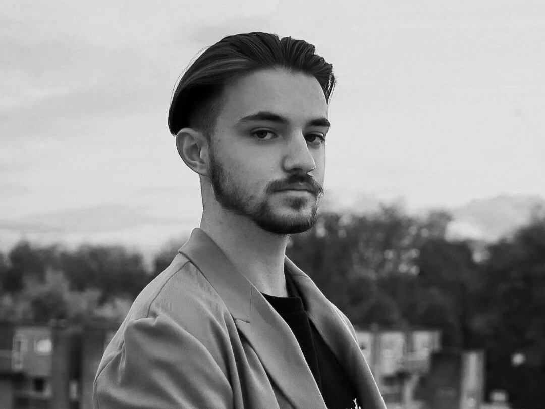 Black and white photo of a young man with styled hair, a beard, and a serious expression standing outdoors with a blurred background of trees and buildings.