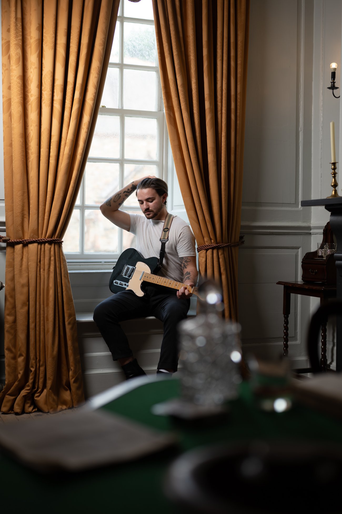 A young man sitting on a window seat with golden curtains playing an electric guitar, with a blurry object in the foreground and a candle holder on a side table.
