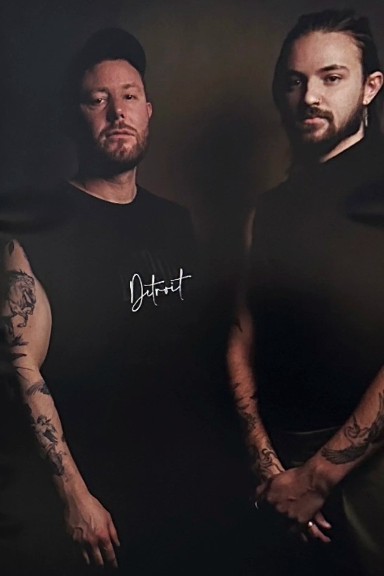 Two men with tattoos wearing black shirts, one with a cap, standing in a dimly lit room with a dark background.
