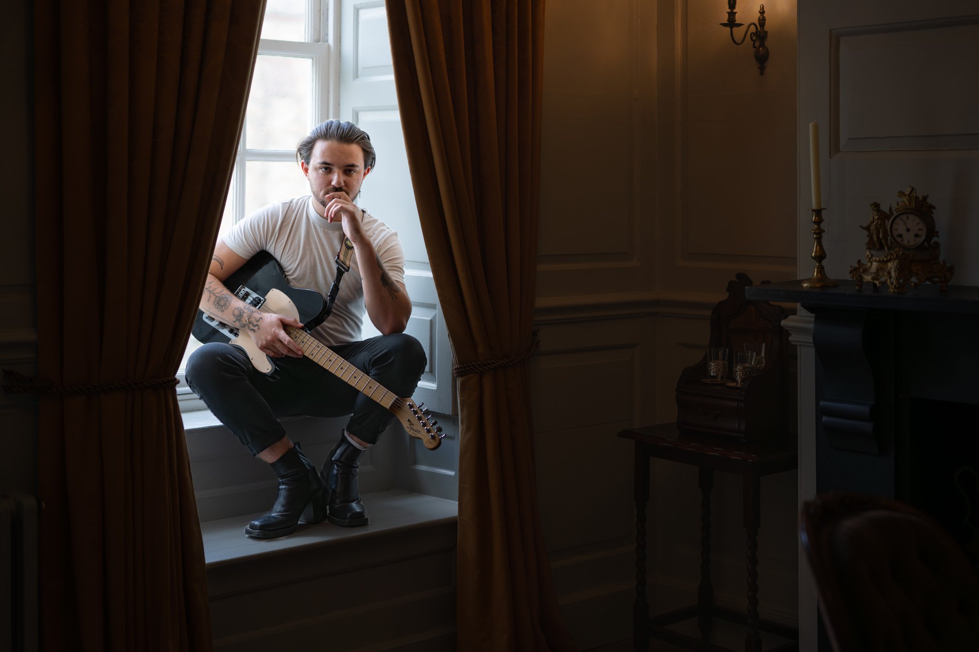 Man sitting on windowsill with guitar, looking at camera in dimly lit room with curtains, candles, and antique clock.