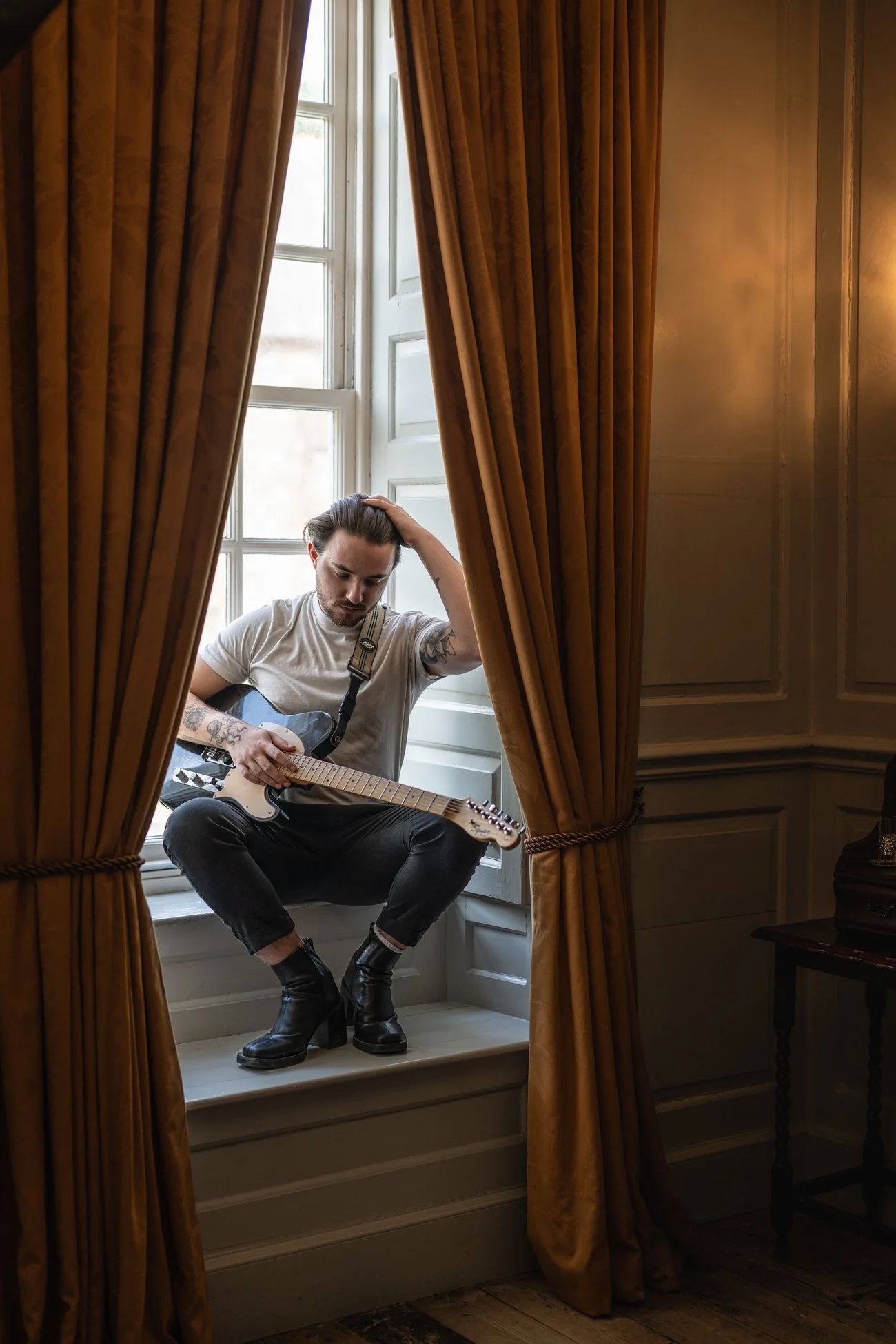 A man with tattoos on his arms, wearing a white t-shirt, black pants, and black boots, is sitting on a window sill playing an electric guitar. He has his head resting on his hand and is surrounded by brown curtains in a well-lit room.