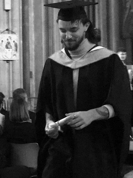 A man in graduation cap and gown holding a diploma, smiling, at a graduation ceremony.