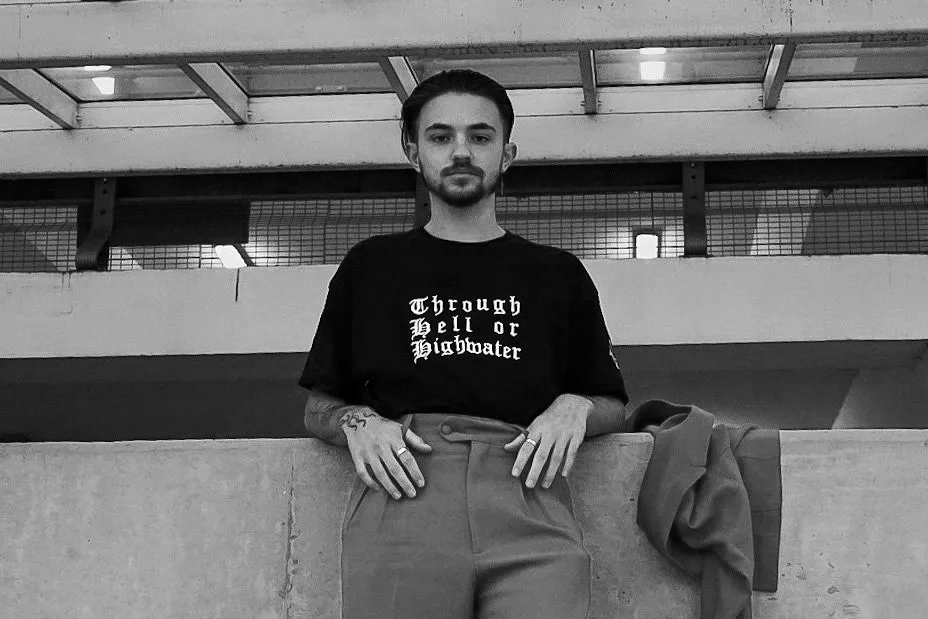 Young man with short dark hair, beard, wearing a black T-shirt with white Gothic text, and high-waisted pants, standing against a concrete wall in an urban environment.