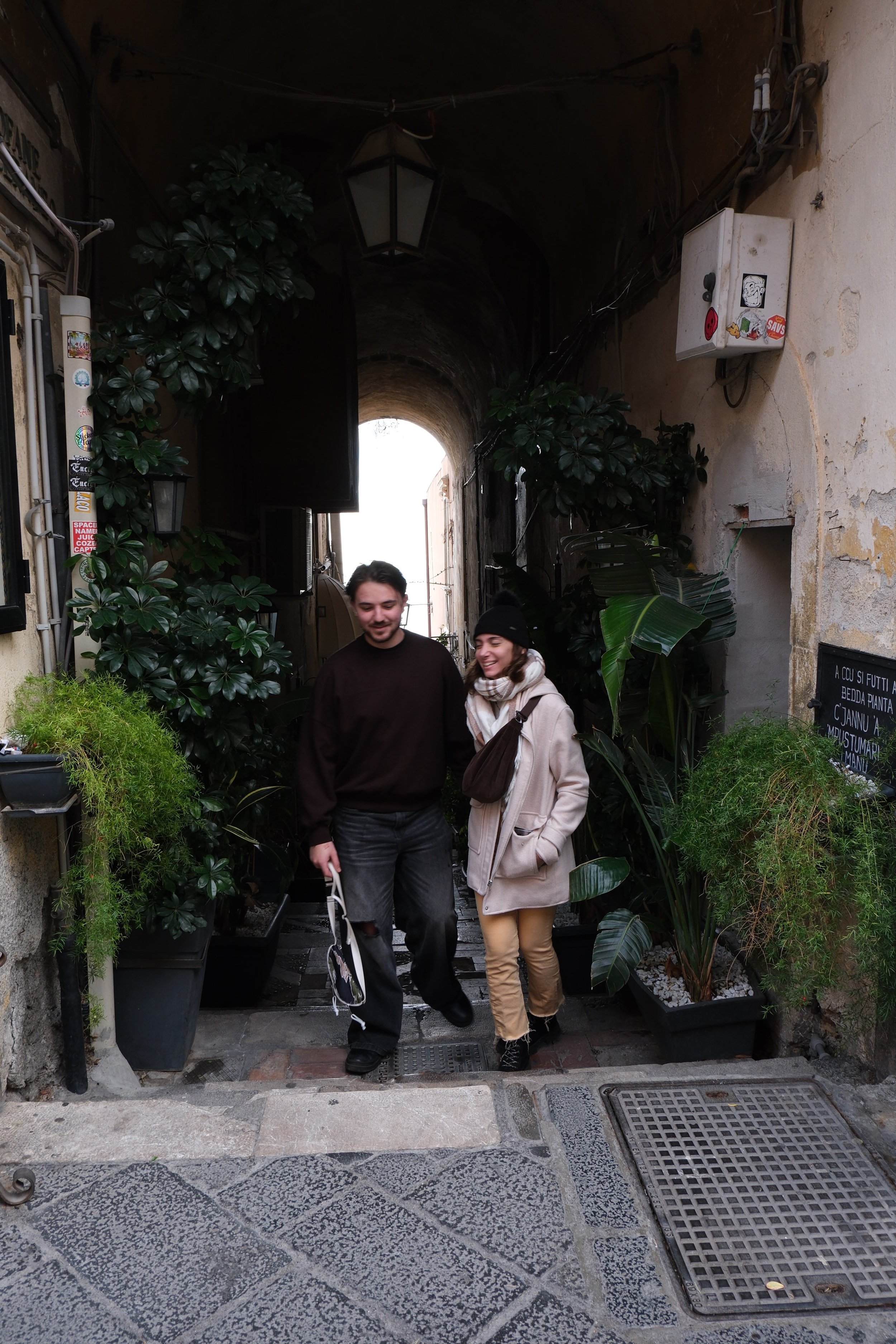 Two people walking through a narrow, archway entrance decorated with green plants and potted foliage, with a street visible in the background.