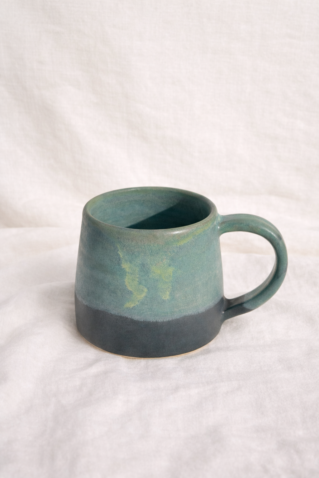 A ceramic mug with a gradient of green and dark gray colors, placed on a white fabric surface.
