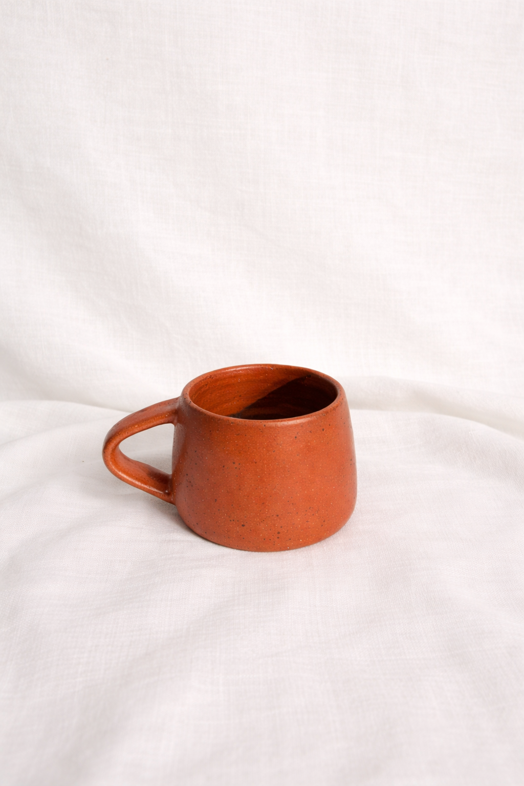A rustic, orange ceramic mug with a handle, placed on a white fabric surface.